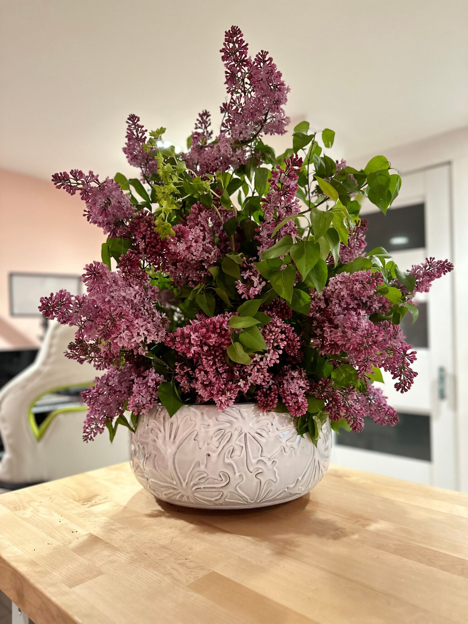 Vase with purple lilac flowers and green leaves on a wooden table in a room.