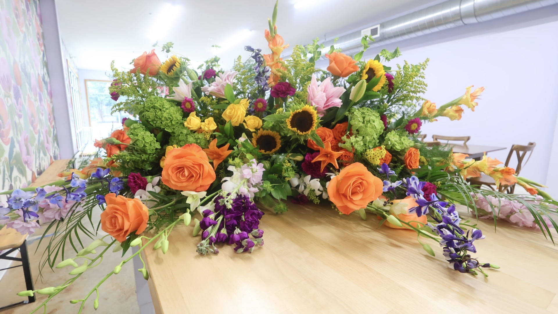 Colorful flower arrangement with roses, sunflowers, and purple flowers on a wooden table in a bright room.
