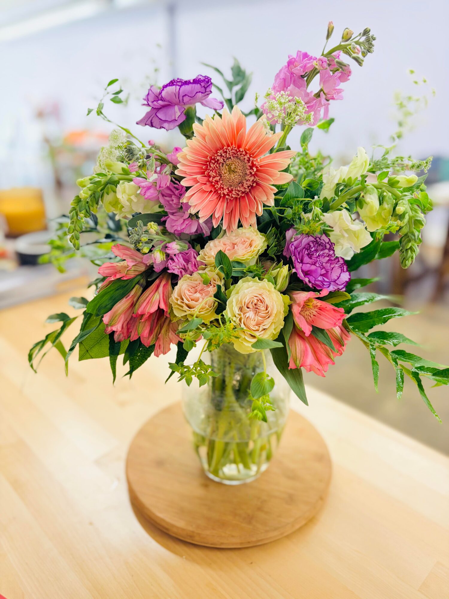 Colorful flower bouquet in a glass vase on a wooden table, with pink, purple, peach, and green flowers and leaves.