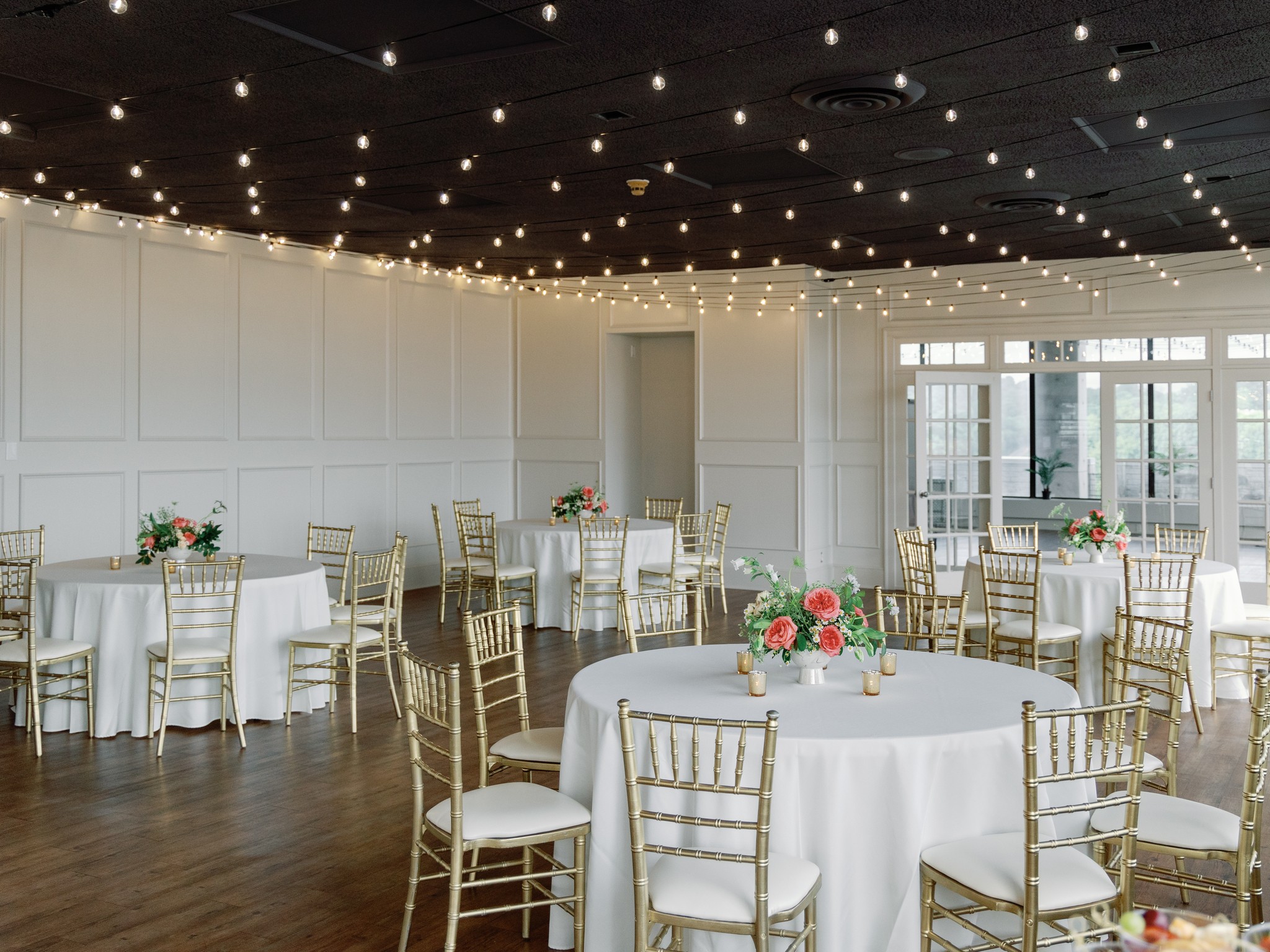 Elegant event space with round tables, white tablecloths, floral centerpieces, gold chairs, and string lights on dark ceiling.