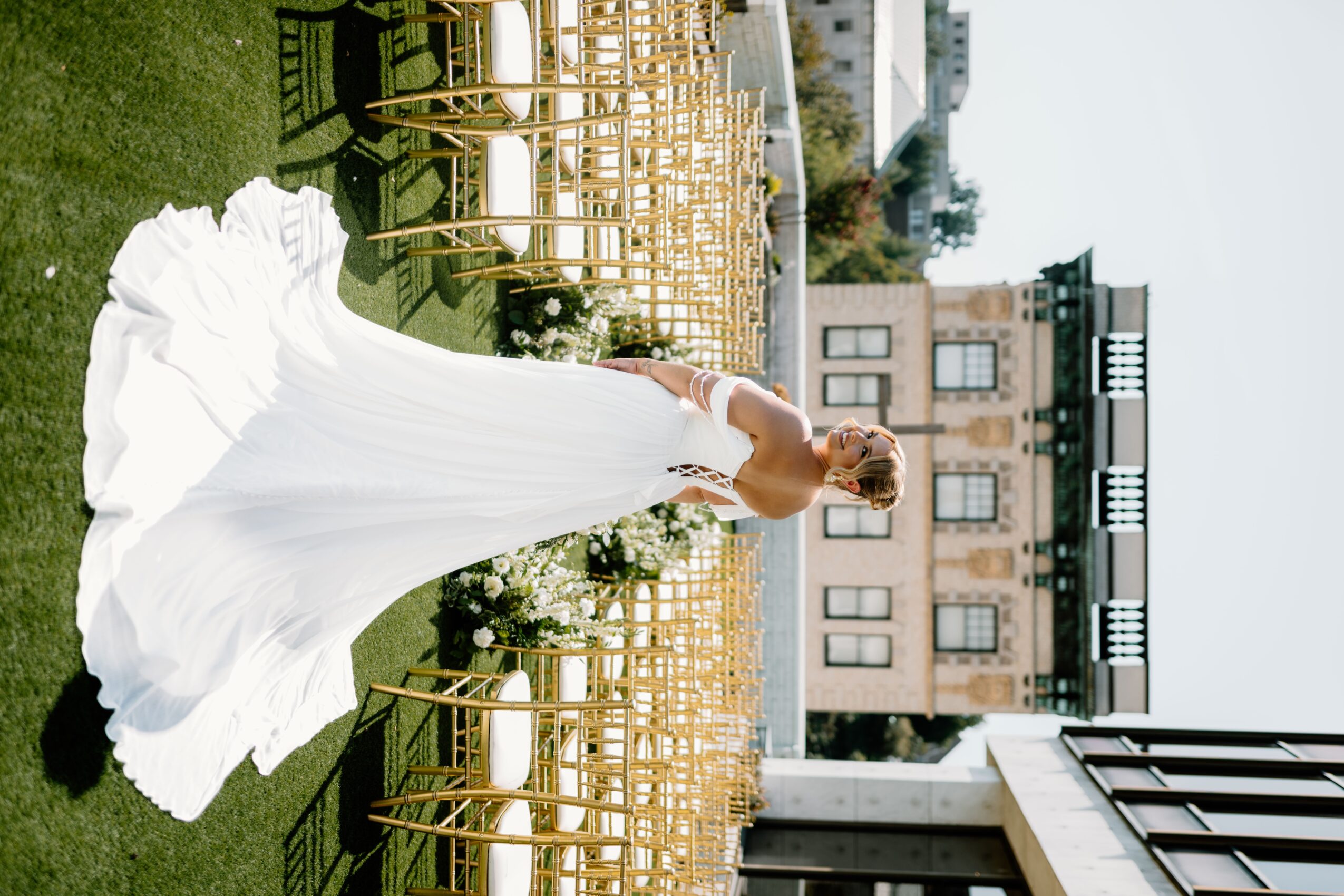 Woman in a white dress standing outdoors with chairs and a building in the background.
