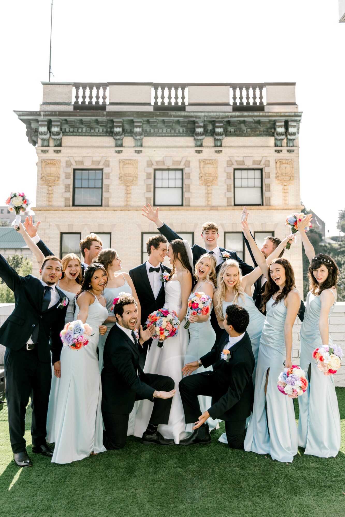 Group of wedding party members outdoors in front of a historic building, holding bouquets, celebrating, smiling, and raising hands.