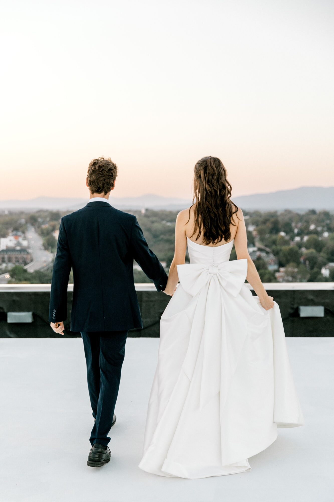 A couple stands on a rooftop, holding hands, with a cityscape and sky in the background.