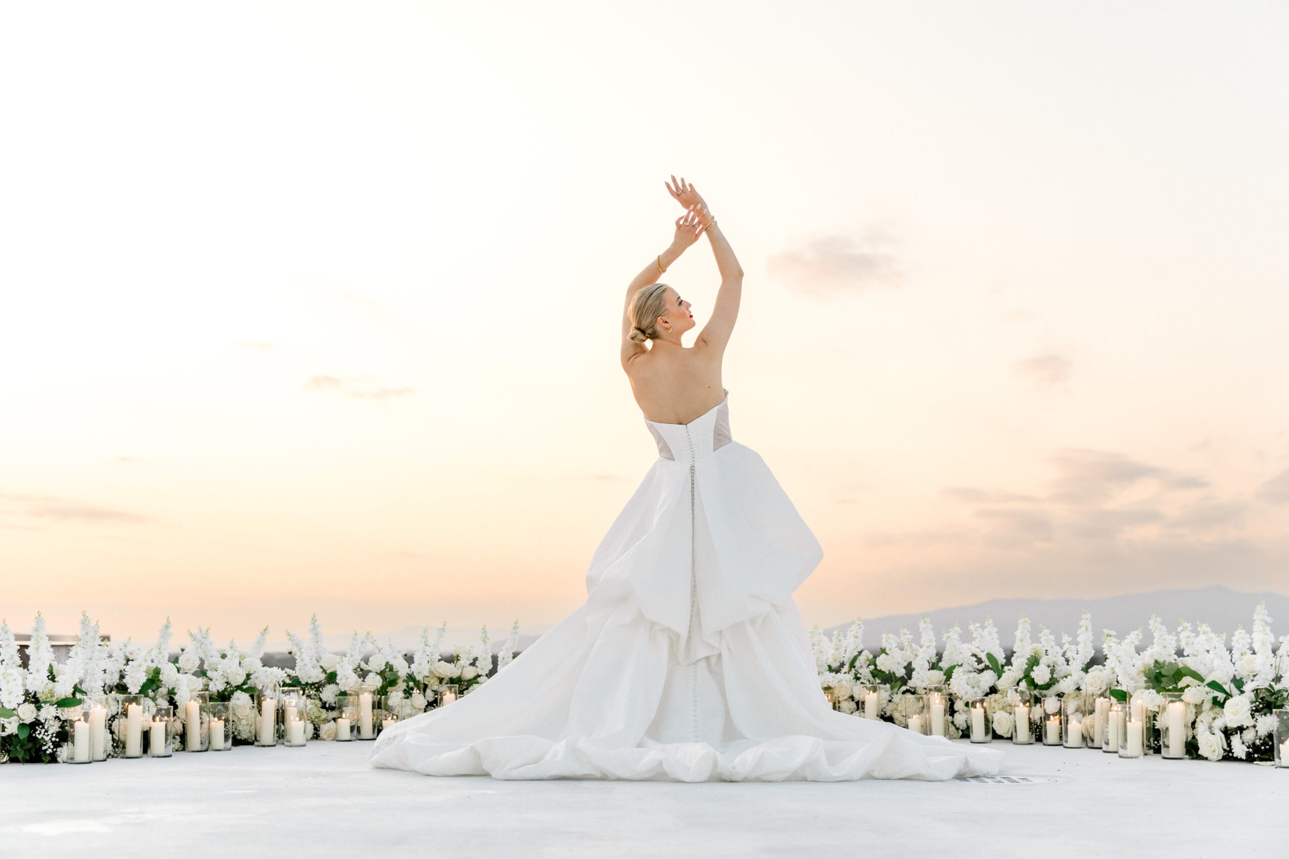 Person in a white dress standing outdoors with arms raised, surrounded by flowers, during sunset or sunrise.