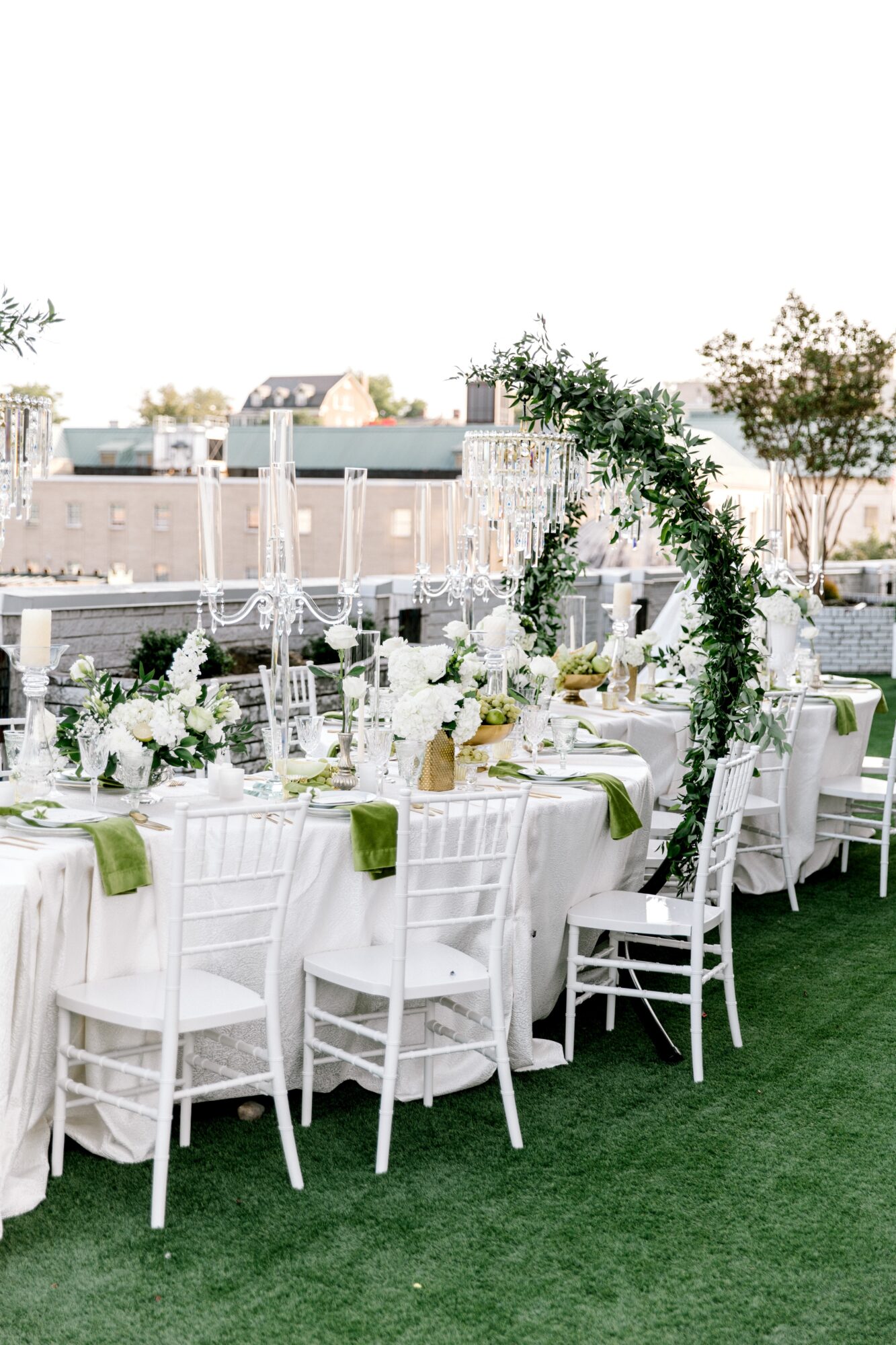 Outdoor event setup with round tables, white chairs, floral centerpieces, and a green arch, on a grassy area.