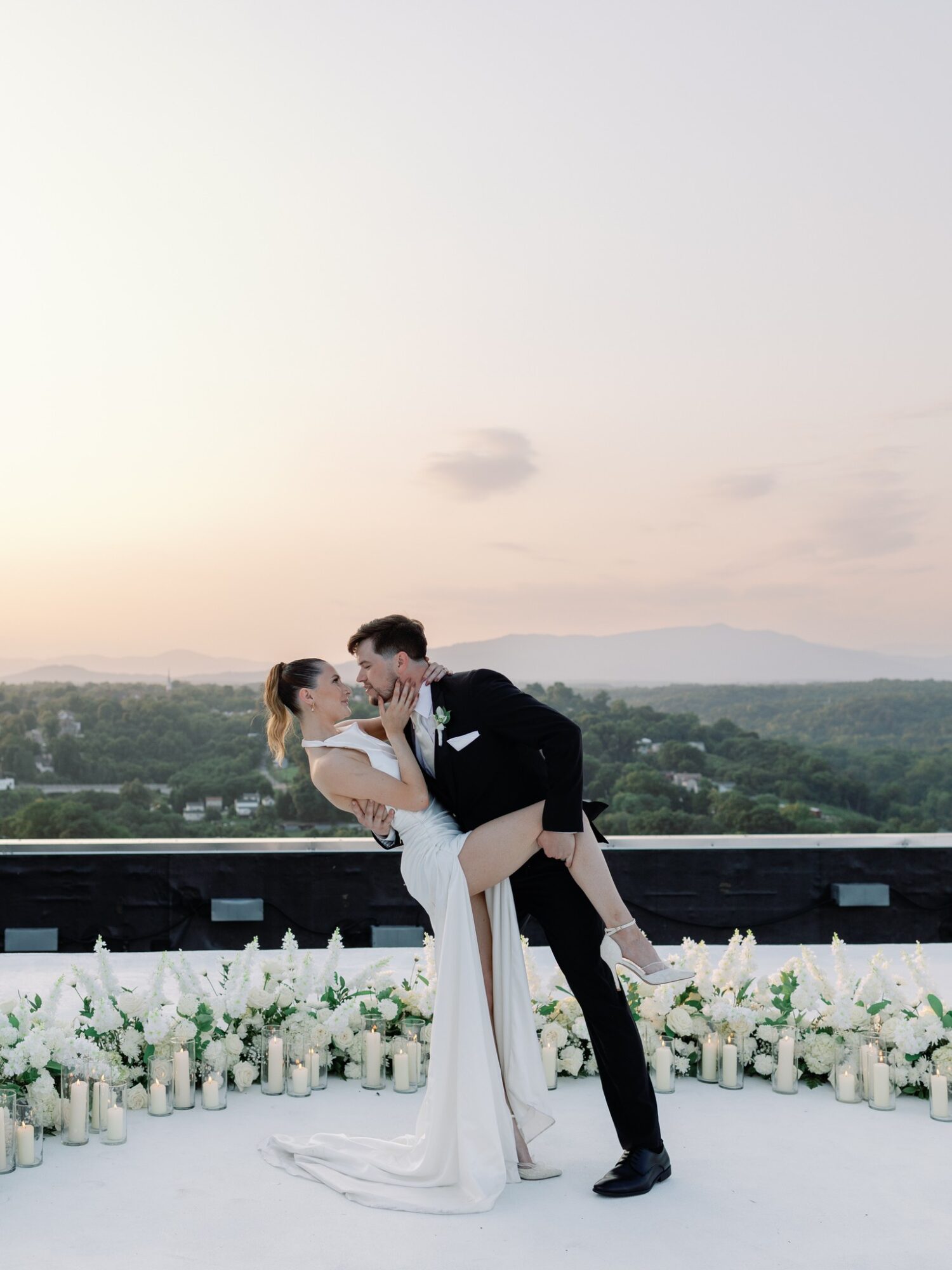 A couple in wedding attire on a balcony with a cityscape and sky in the background, the groom dips the bride.
