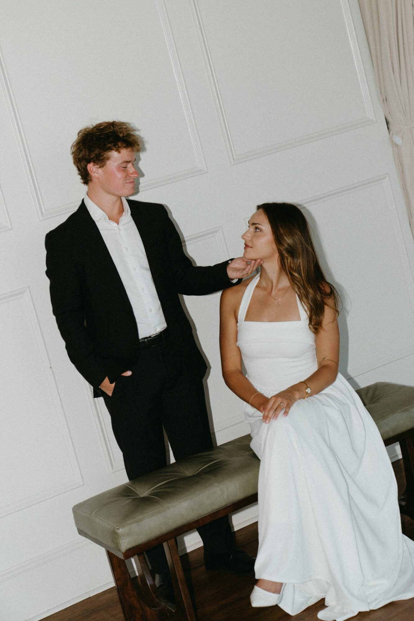 A man in a black suit and a woman in a white dress are indoors, with the man standing and the woman sitting on a bench.