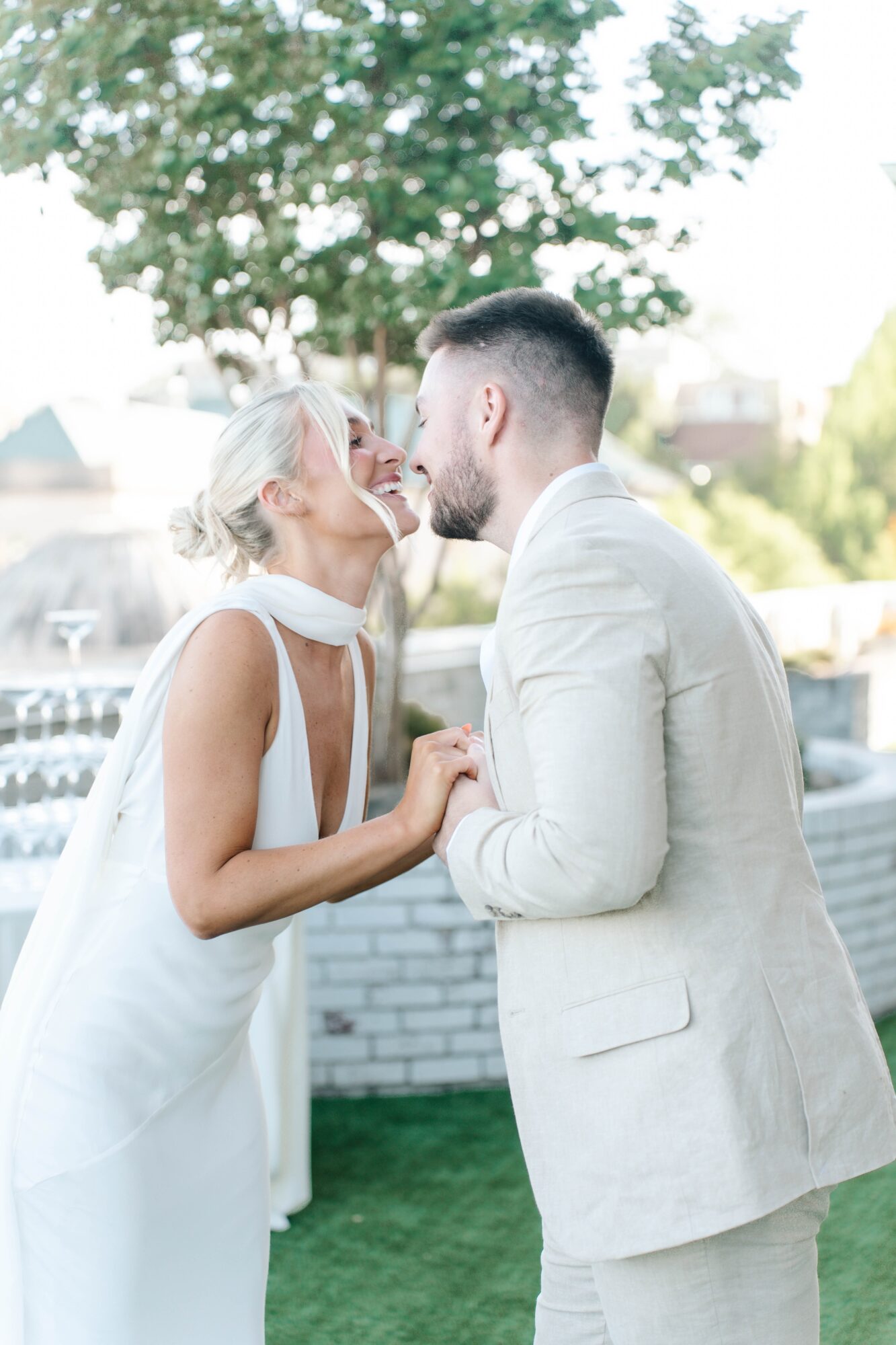 A bride and groom lean in close, smiling, outdoors with trees and a building in the background.