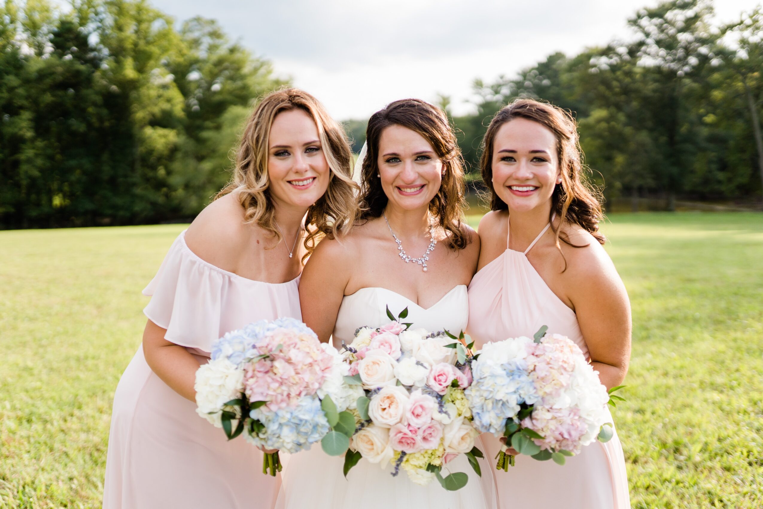 Three women in white dresses holding bouquets, smiling outdoors on a sunny day with trees in the background.