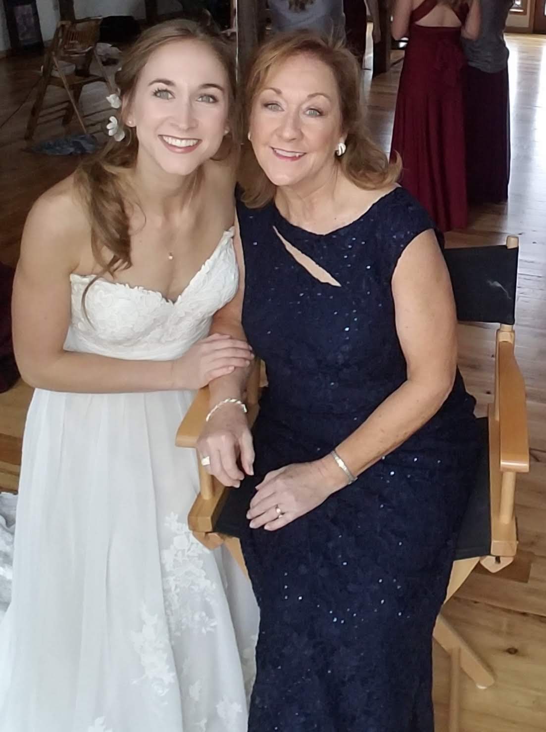 Two women smiling, one in a white dress and the other in a dark blue dress, sitting close together indoors.
