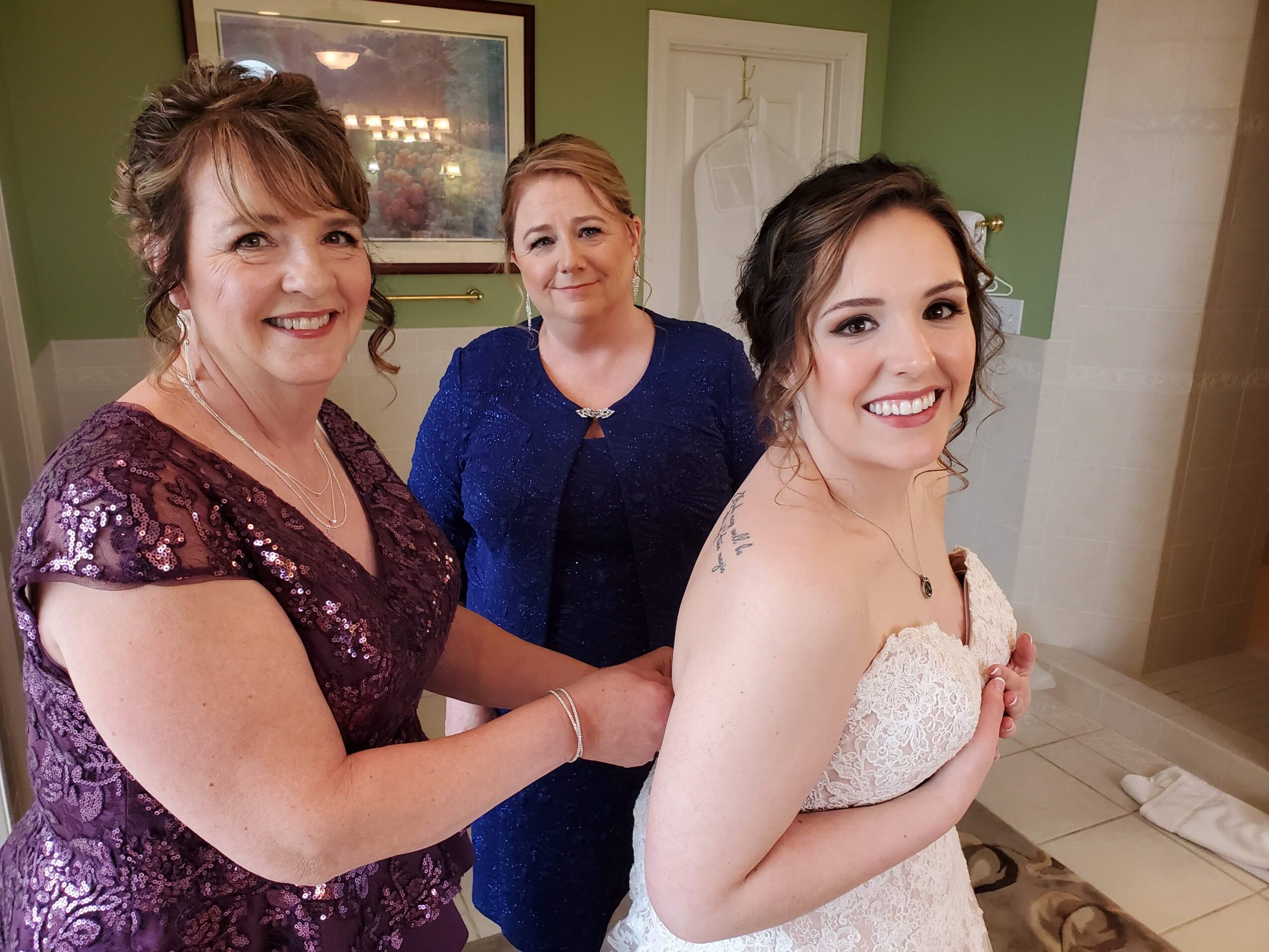 Three women smiling indoors, one in a white dress, two in blue dresses, standing close together.