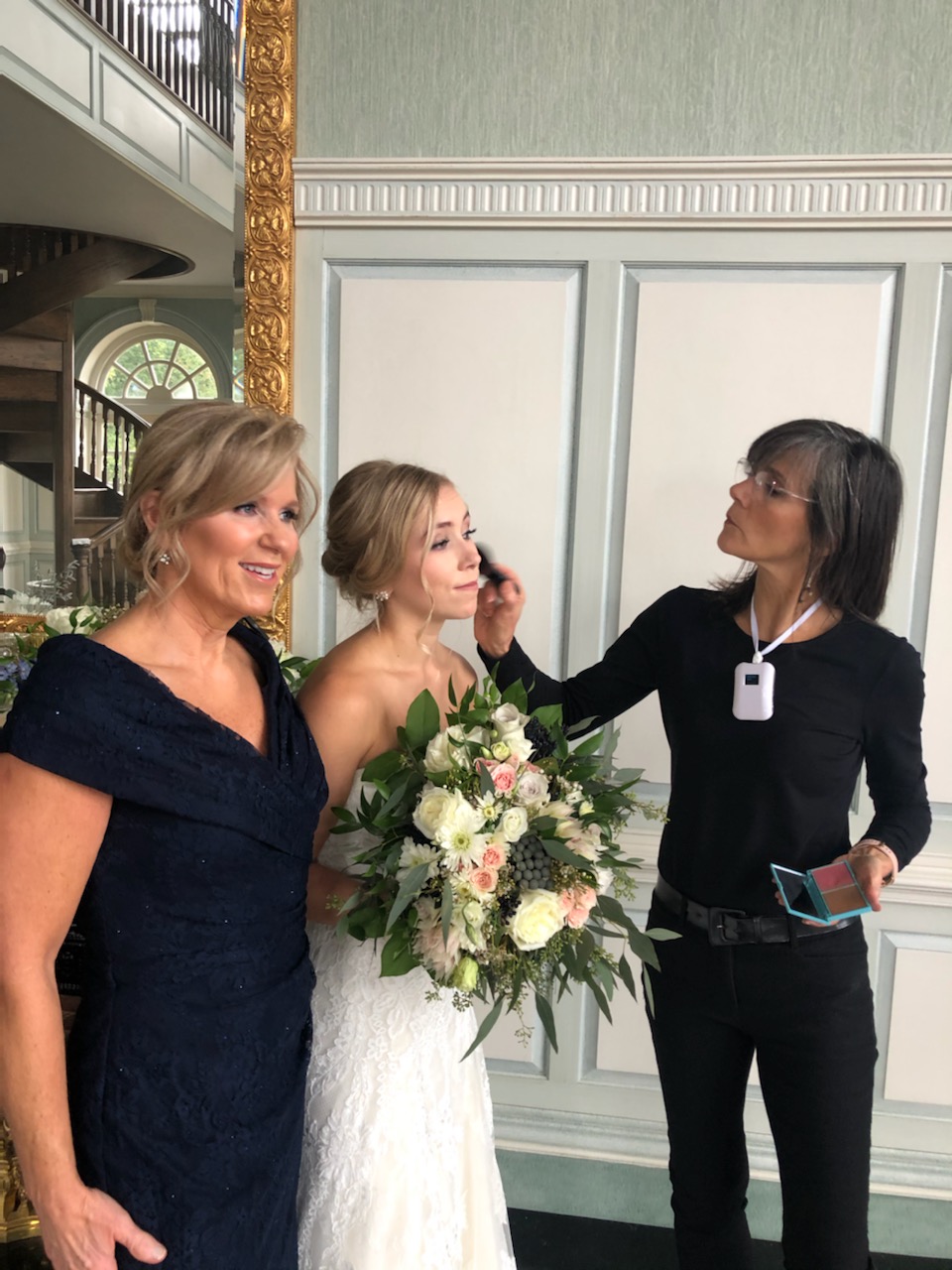 Three women standing indoors, one in a dark dress, one in a wedding gown holding a bouquet, and a woman in black applying makeup.