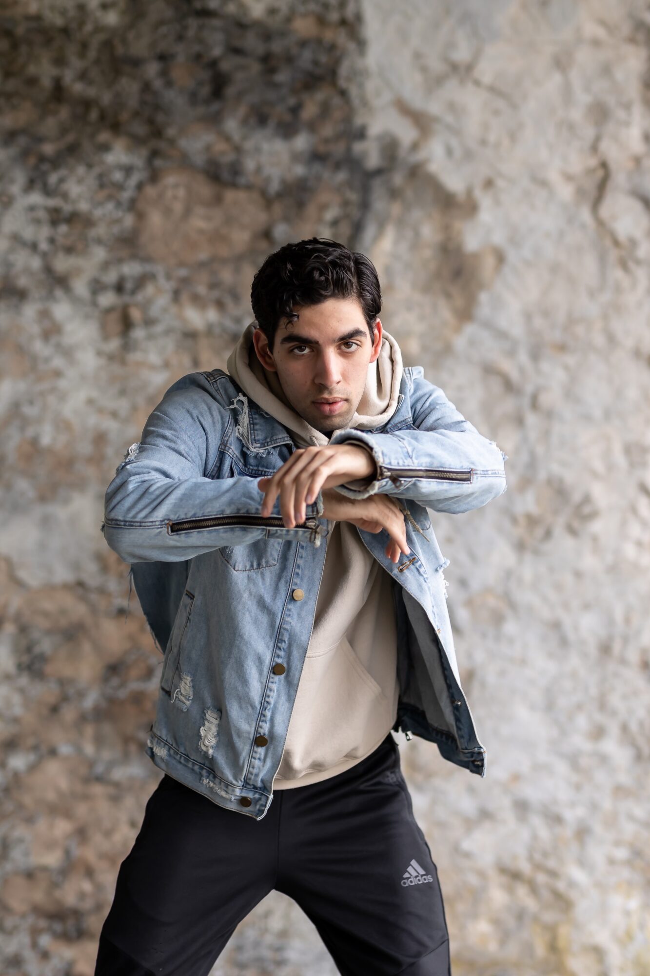 Young man with dark hair wearing a denim jacket and white shirt, standing against a textured stone wall, looking at camera.