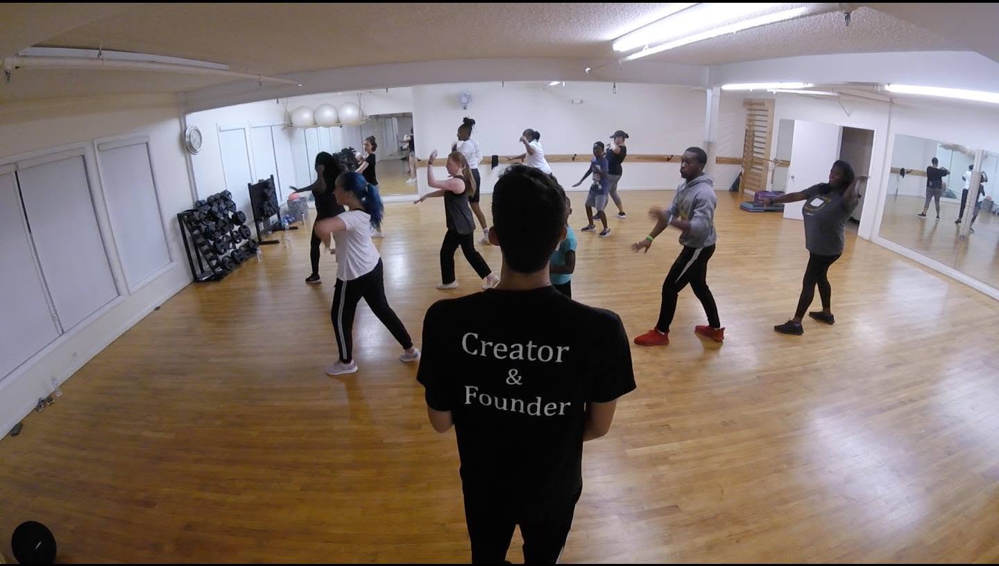 Group of people dancing in a spacious room with wooden floors and mirrors, instructor in foreground wearing a black shirt.