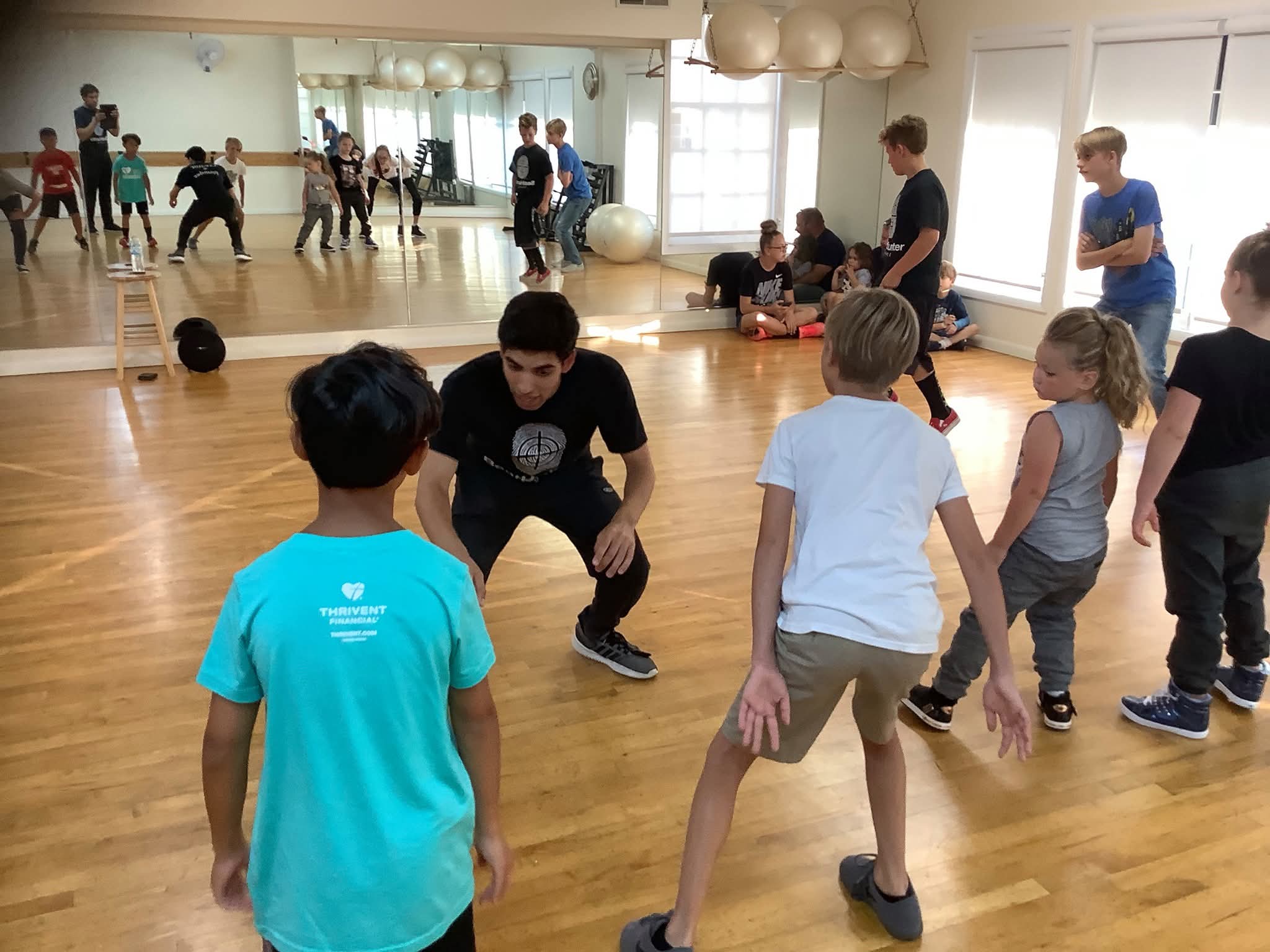 Children and adults in a gymnasium, some playing and others watching, with a mirror reflecting the scene.