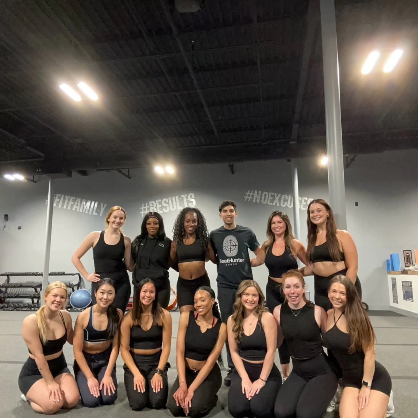 Group of twelve women in black workout clothes posing in a gym, some standing and some kneeling, with gym equipment in background.
