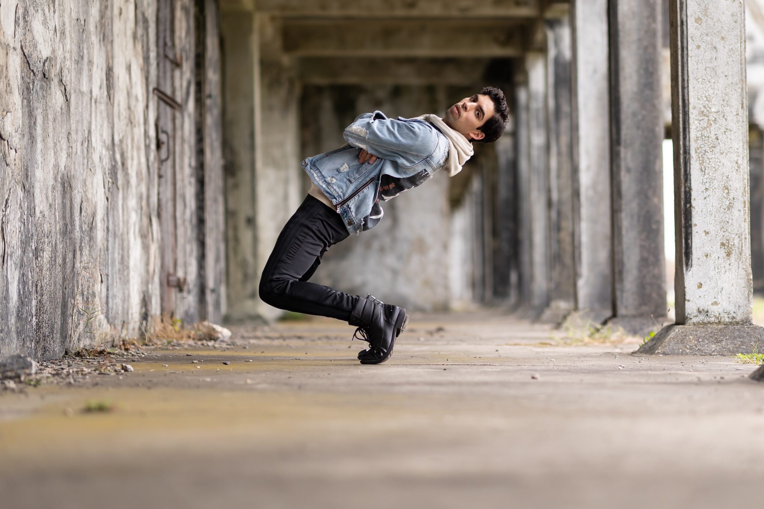 Person leaning backward in an outdoor concrete tunnel, with arms crossed and legs bent, wearing a denim jacket and black pants.