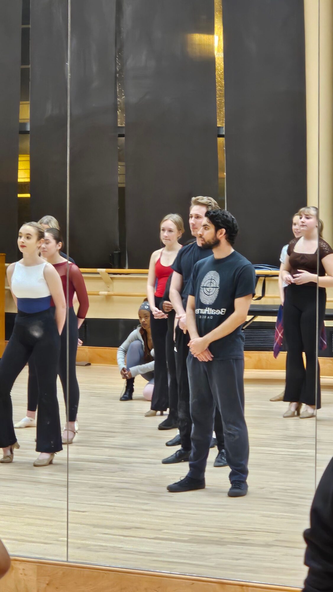 Group of people standing in a dance studio, some holding hands, with a wooden floor and mirror wall.