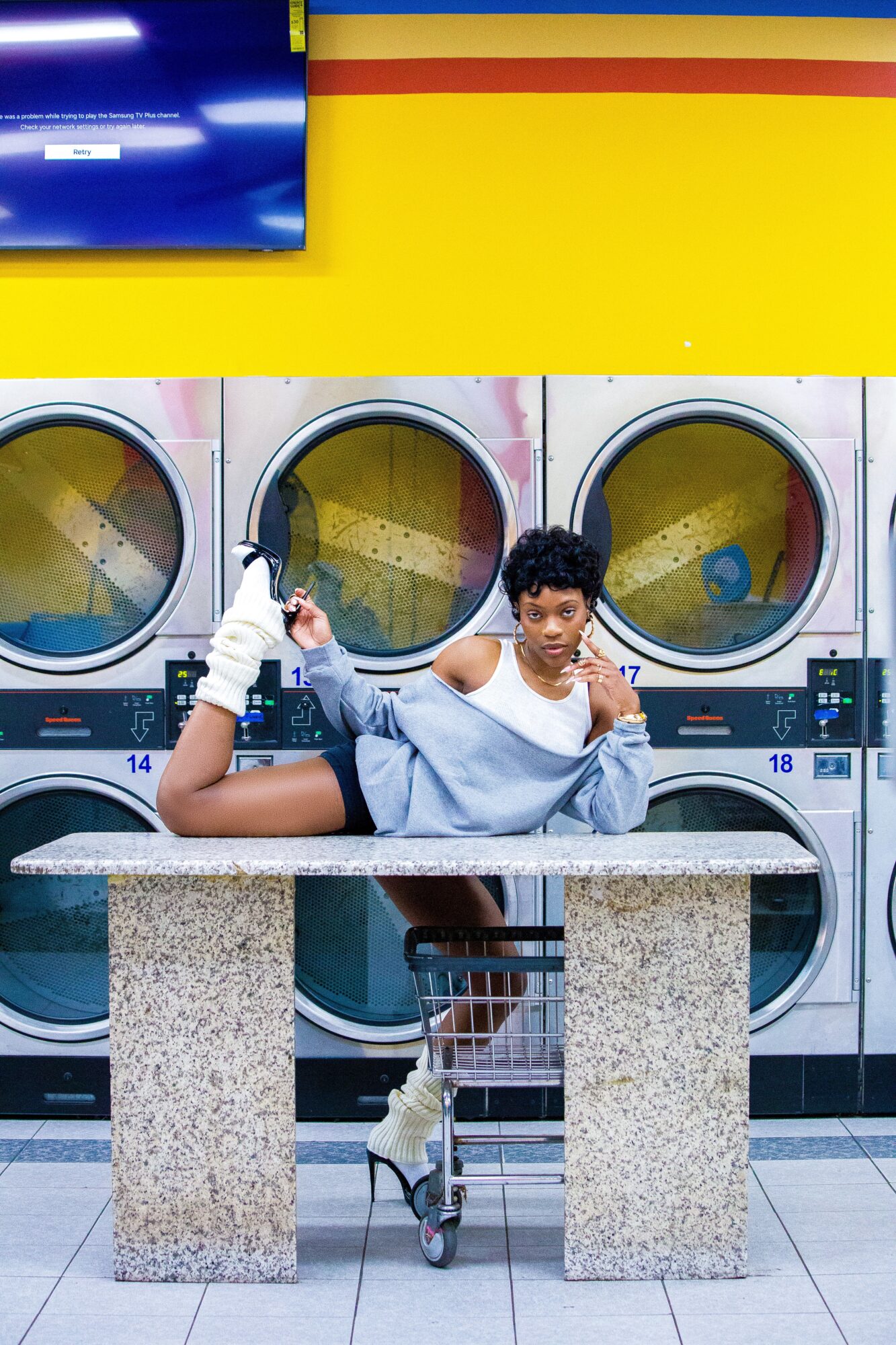 Person with short hair and light clothing lying on a table in laundromat, with washing machines behind.