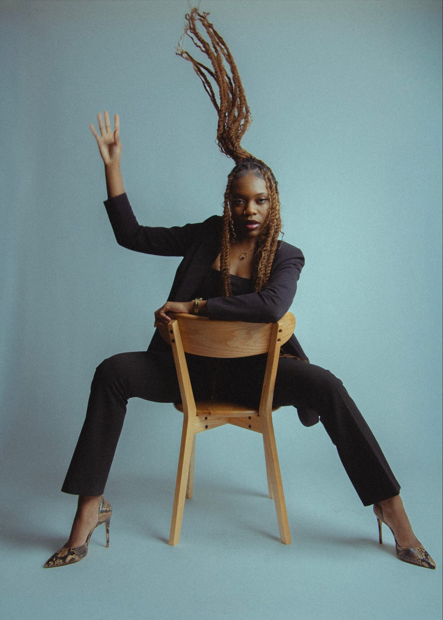 Woman with long braided hair sitting on a chair, legs apart, one arm raised, against a light blue background.