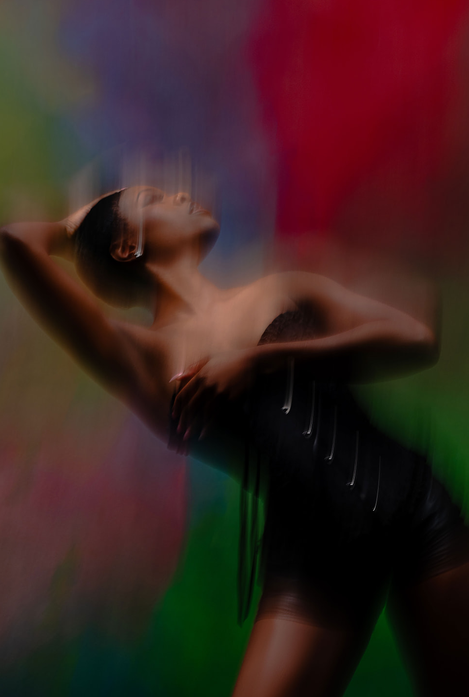 A woman with arms raised, head tilted back, in front of a colorful blurred background, wearing a black outfit.