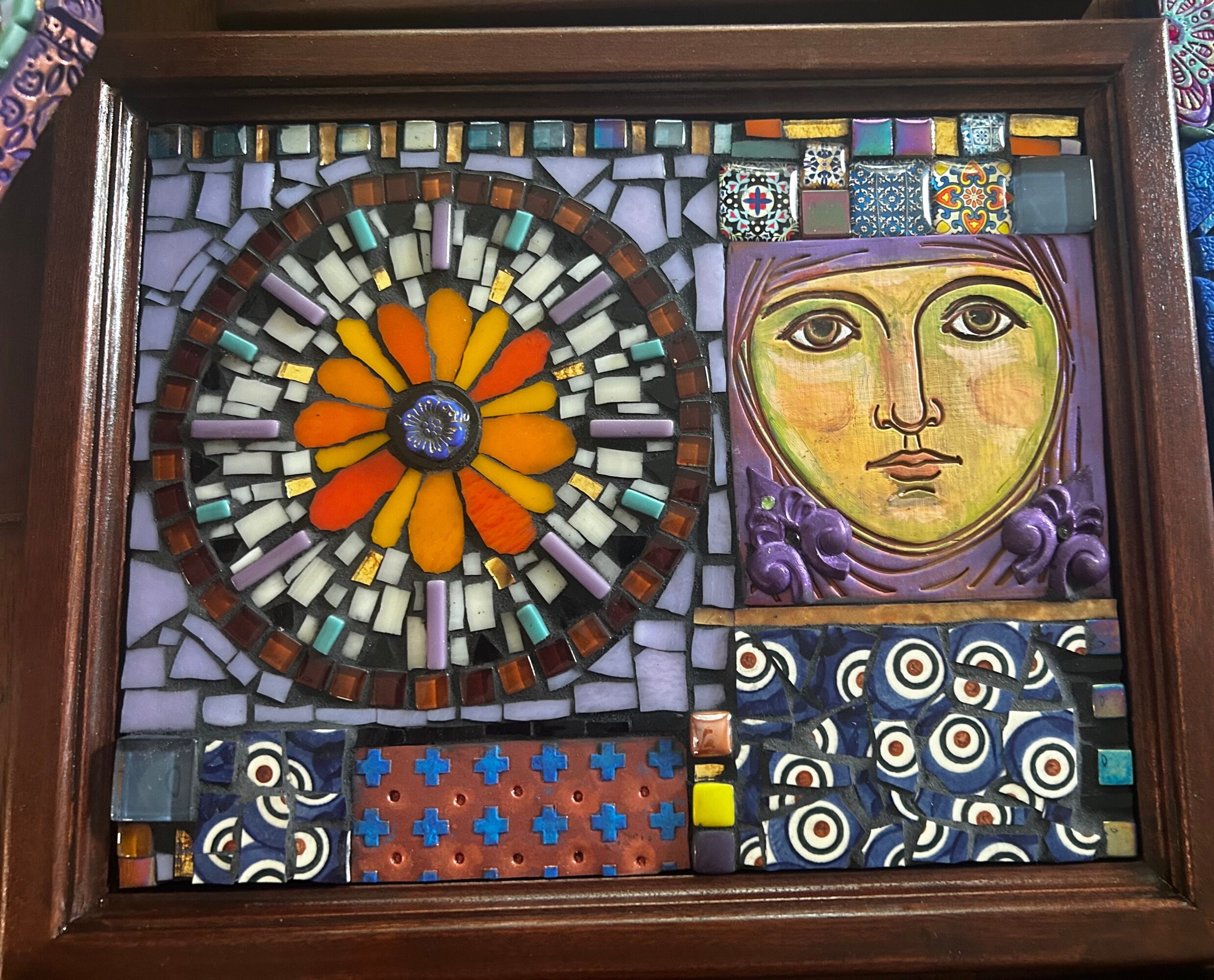 Colorful mosaic with a flower design and a face portrait, framed in wood.