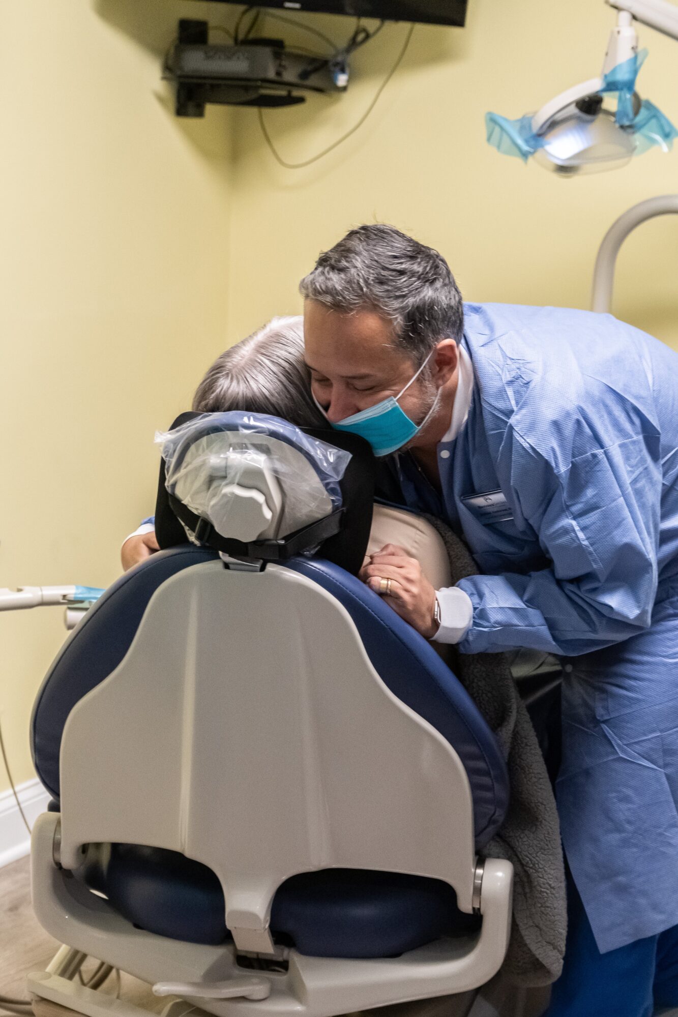 A healthcare professional wearing a blue gown and face mask interacts with a young patient in a dental chair, in a clinical setting.