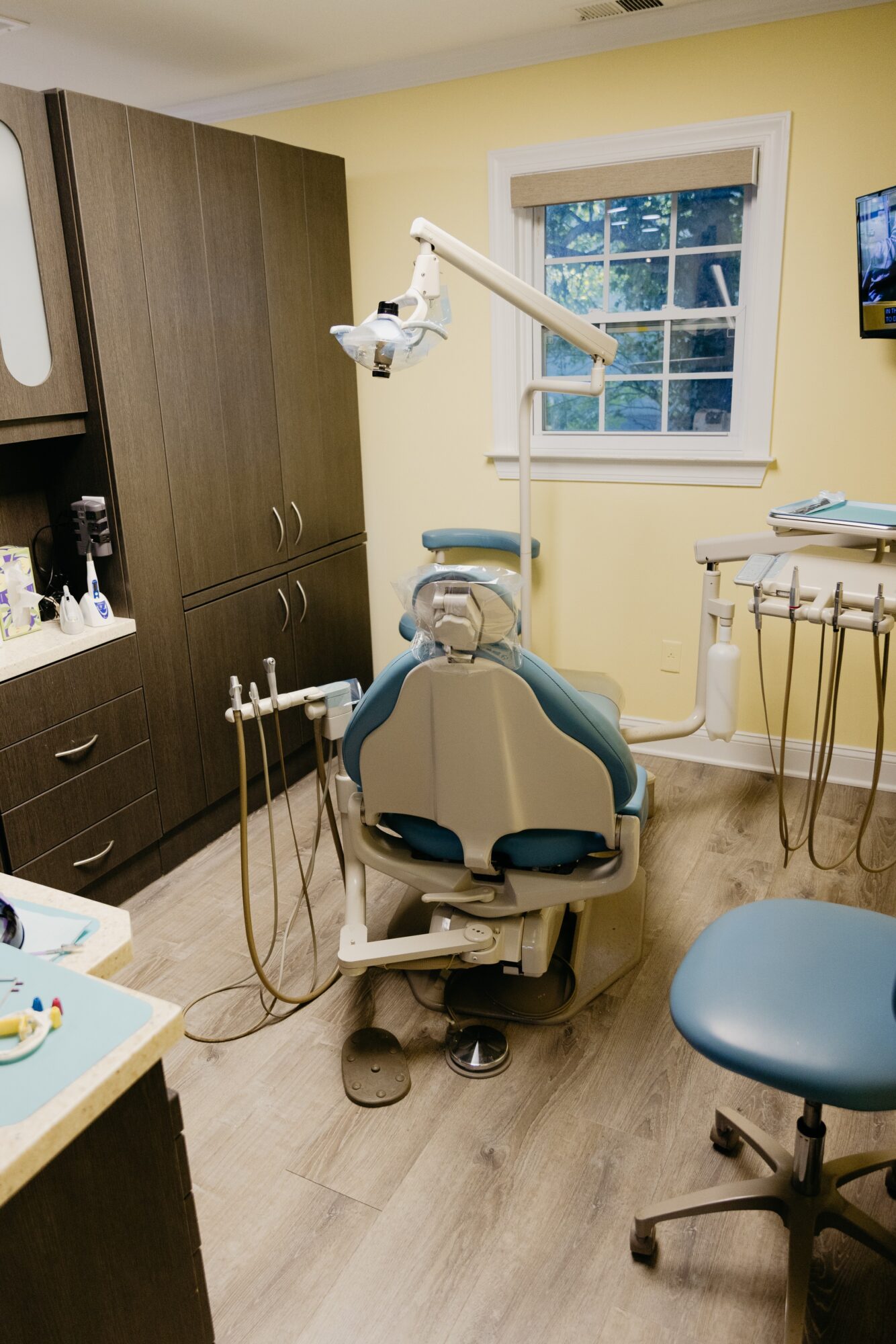 Dental chair in a clinic room with equipment, window, and cabinets, viewed from the front.