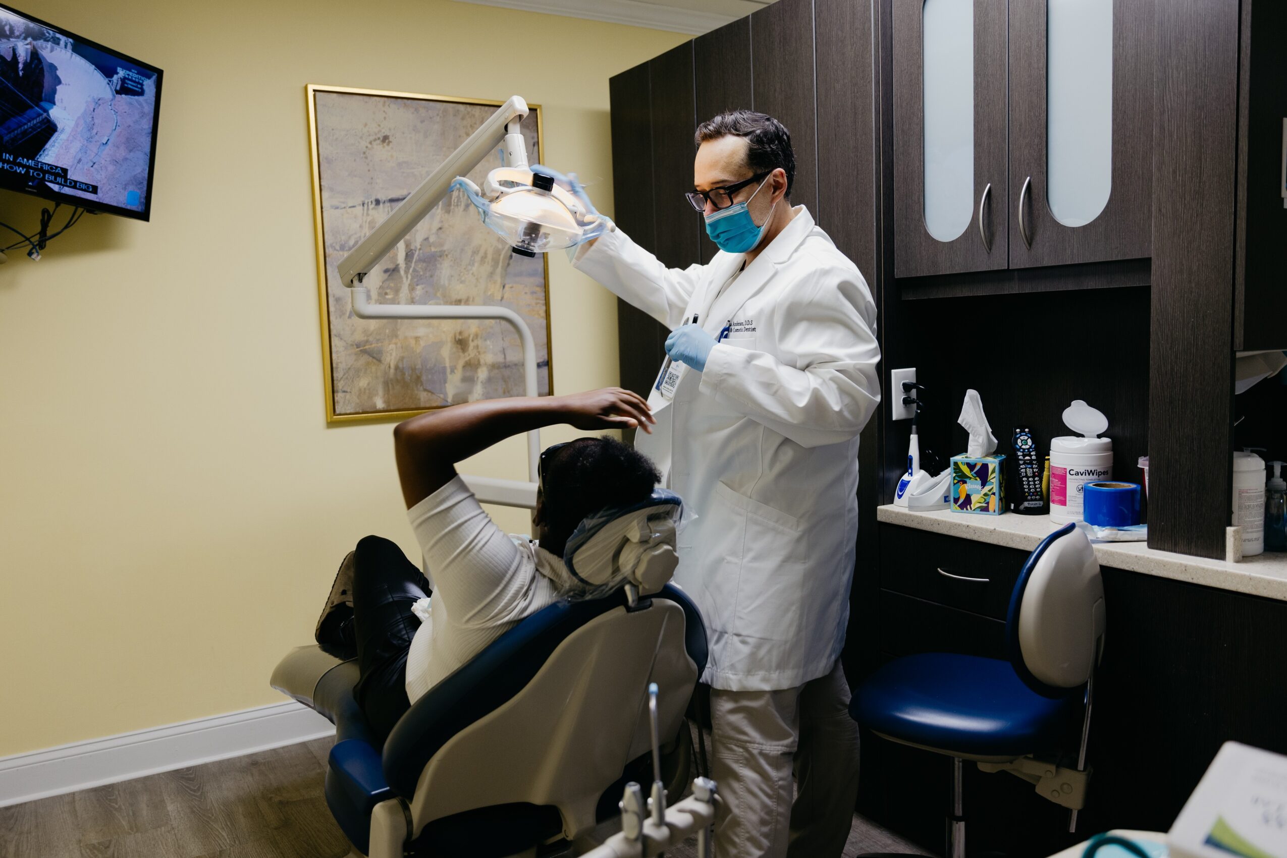 Dentist in white coat and mask examines patient's mouth in dental office, with dental tools and cabinets nearby.