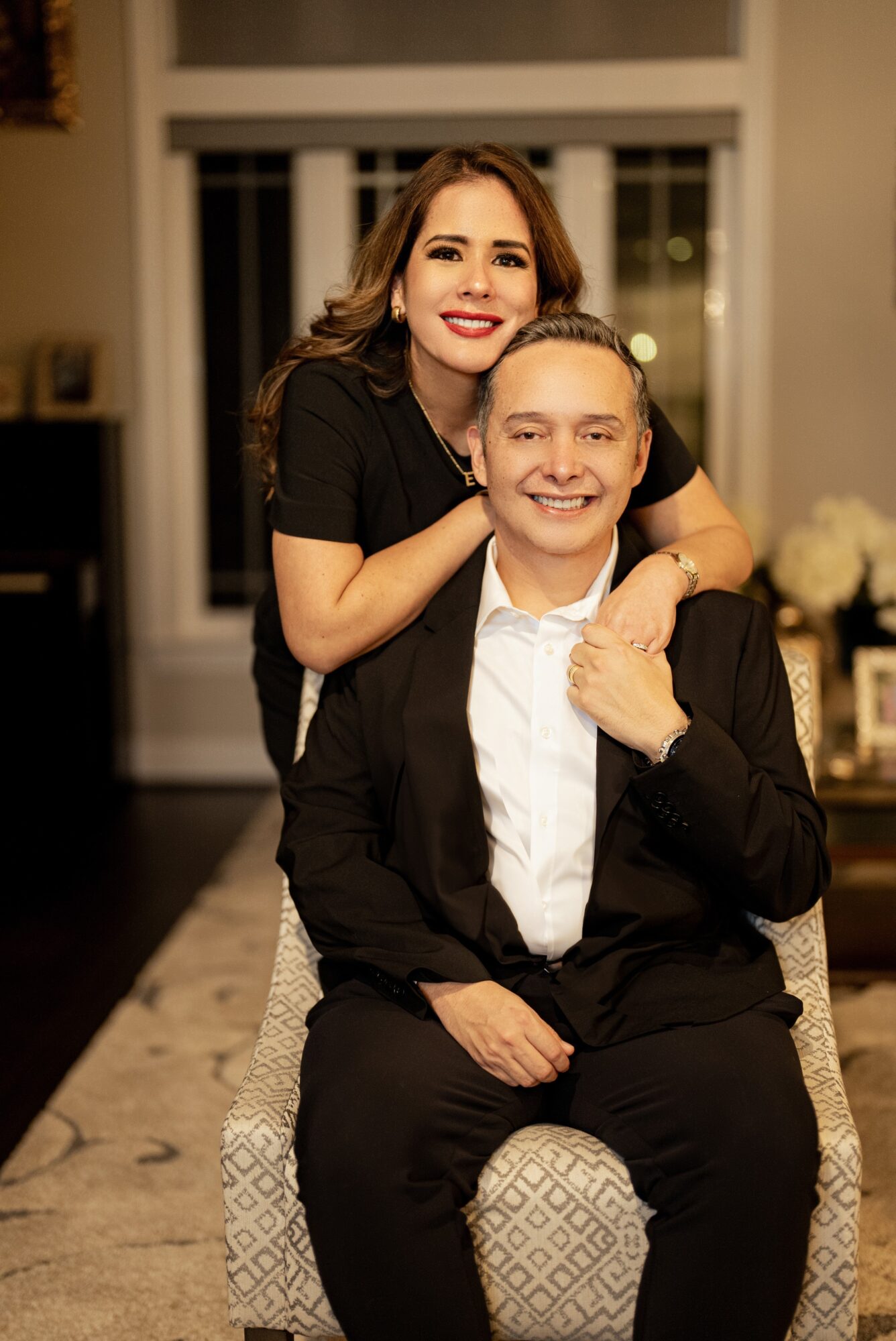 A smiling woman with long brown hair stands behind a seated man, both dressed in formal attire, in a well-lit room.