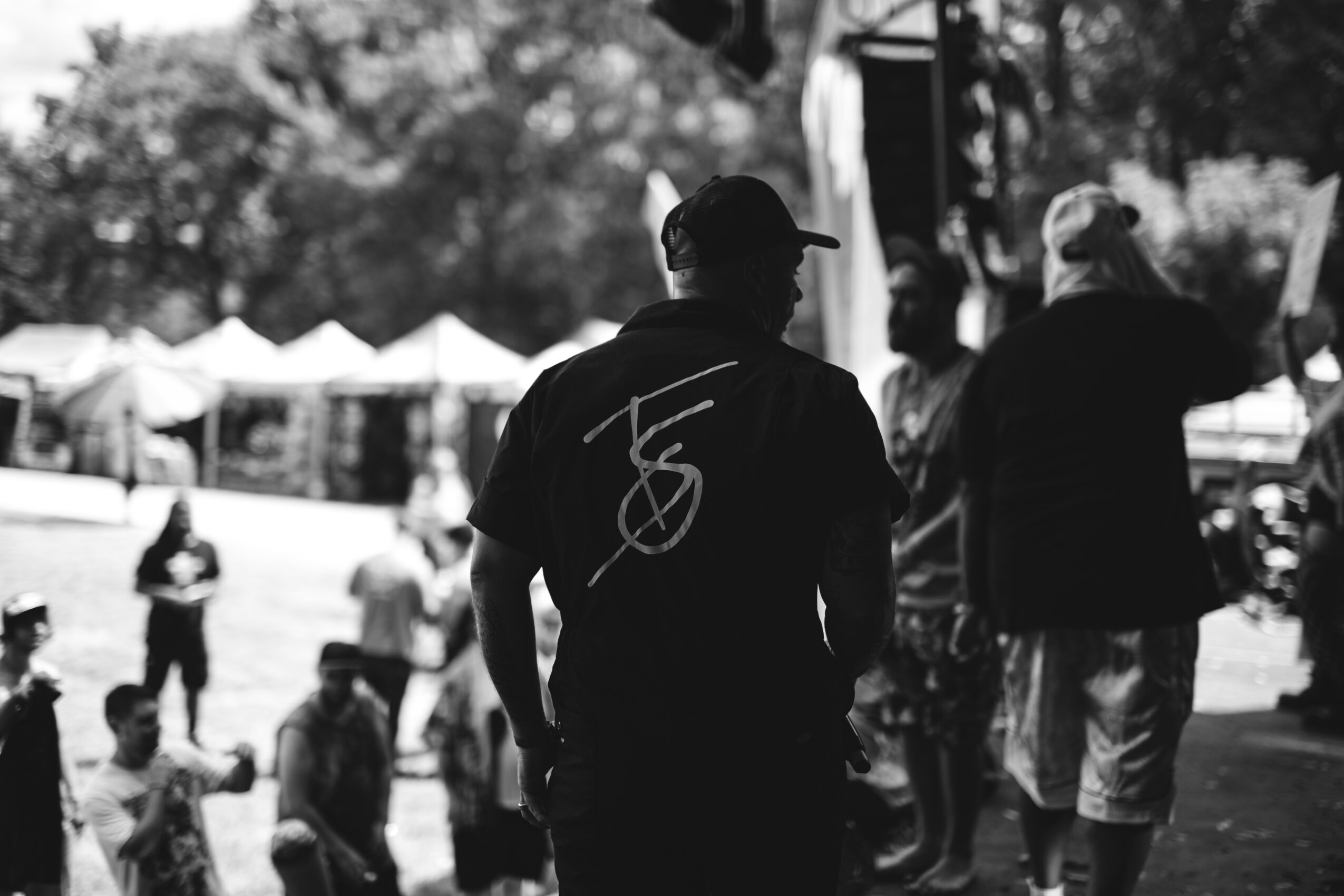 Person wearing a cap and T-shirt with a design, standing outdoors among people and tents.
