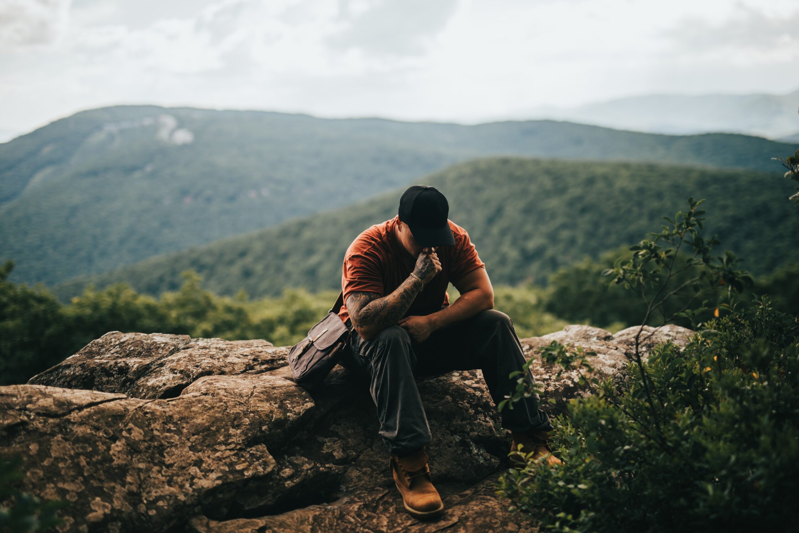 Person sitting on rocks outdoors with mountains and cloudy sky in background, head bowed and hand on face, wearing a red shirt.