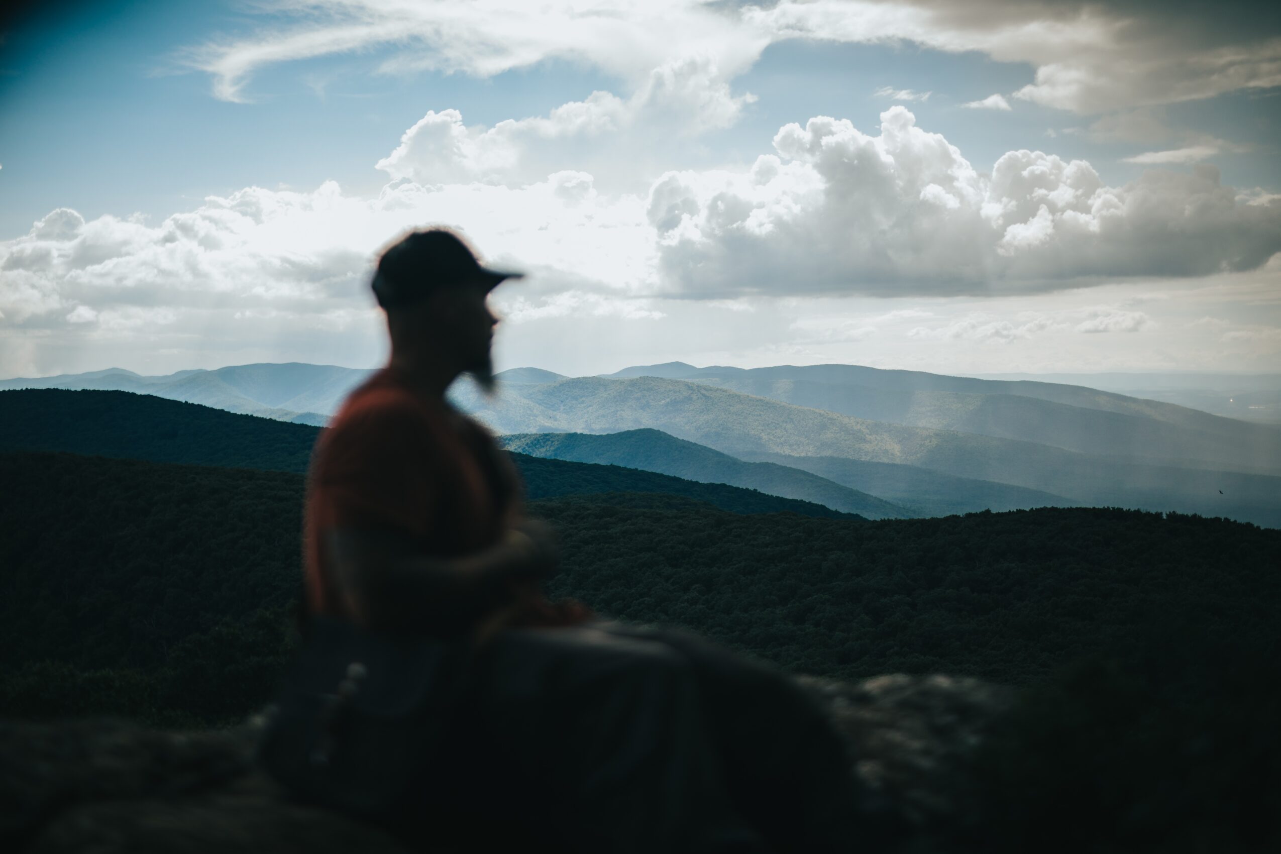 Silhouette of a person sitting outdoors with mountains and cloudy sky in background.