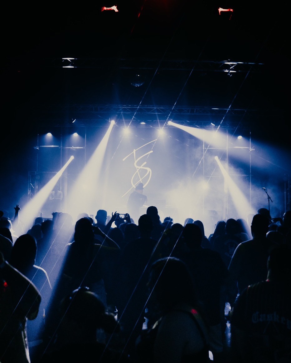 Crowd watching a stage with bright lights and a logo, dark background, silhouettes of people, concert or event setting.
