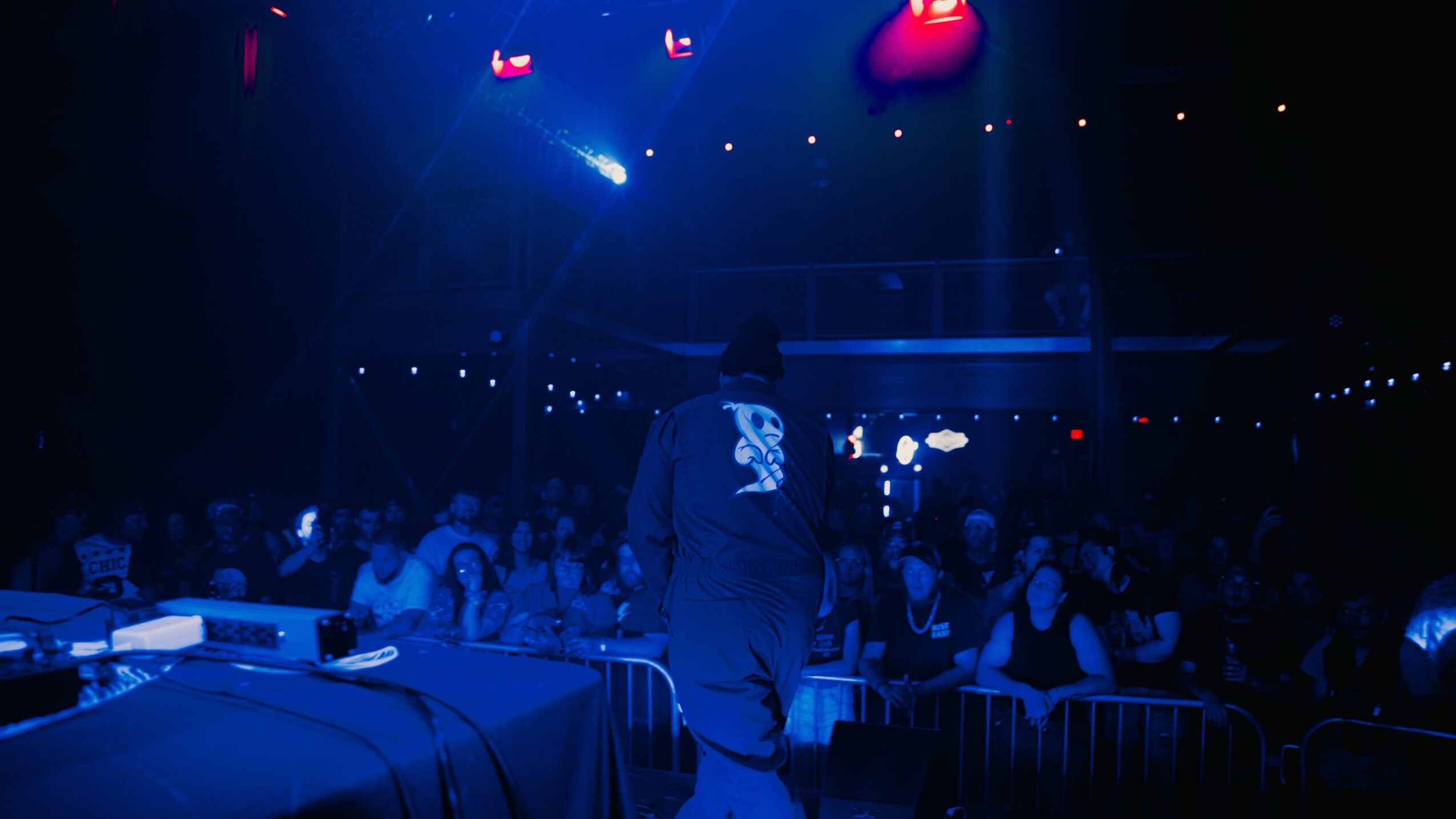 Person on stage with DJ equipment in front, audience watching in a dark, colorful concert venue.