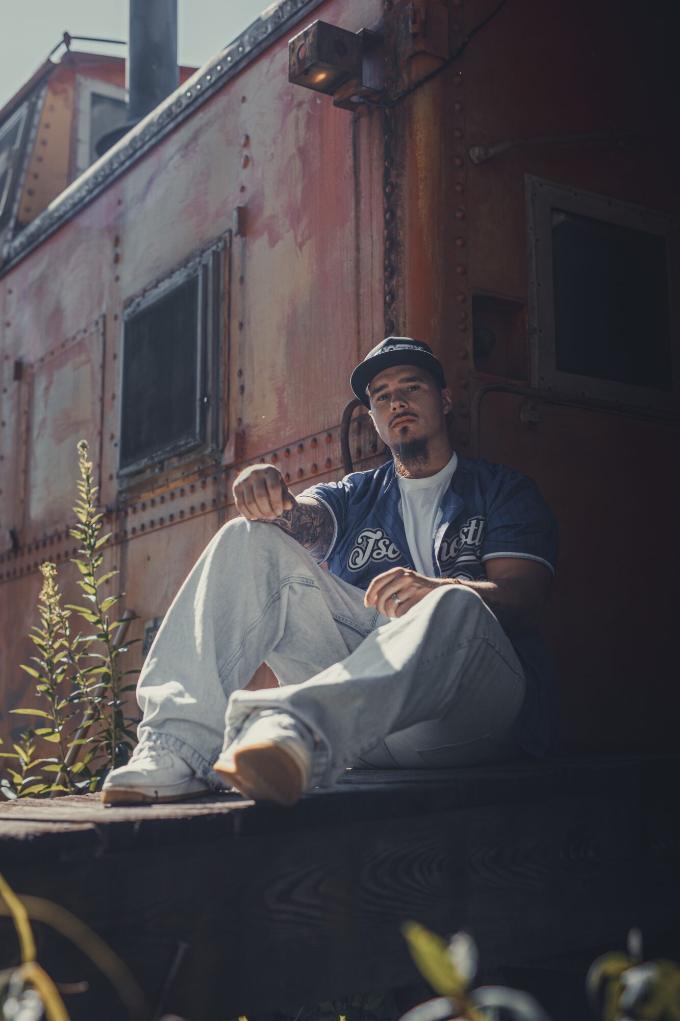 Young man sitting outdoors against a rusty metal wall, wearing a cap, jacket, and white pants, with plants nearby.