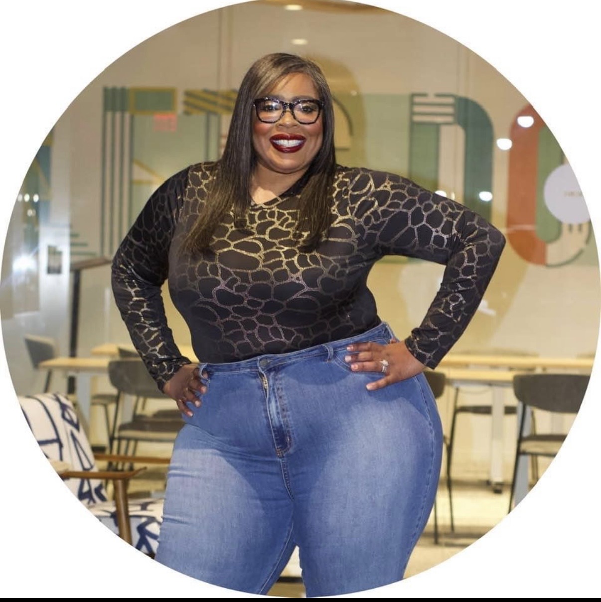 Woman with glasses smiling, wearing a black patterned top and blue jeans, standing indoors with chairs and artwork behind her.