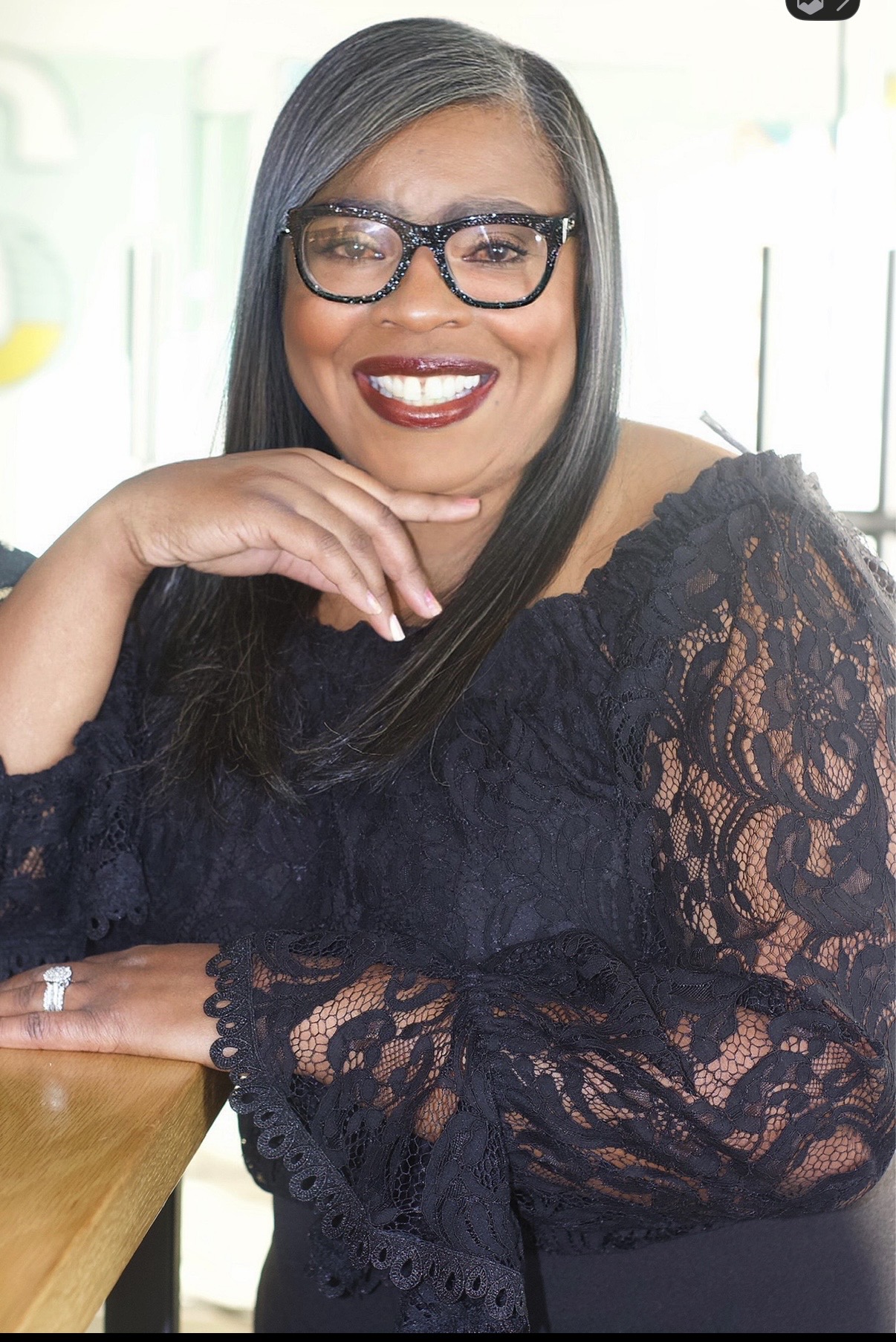 Smiling woman with glasses and long dark hair, wearing a black lace top, resting her chin on her hand.