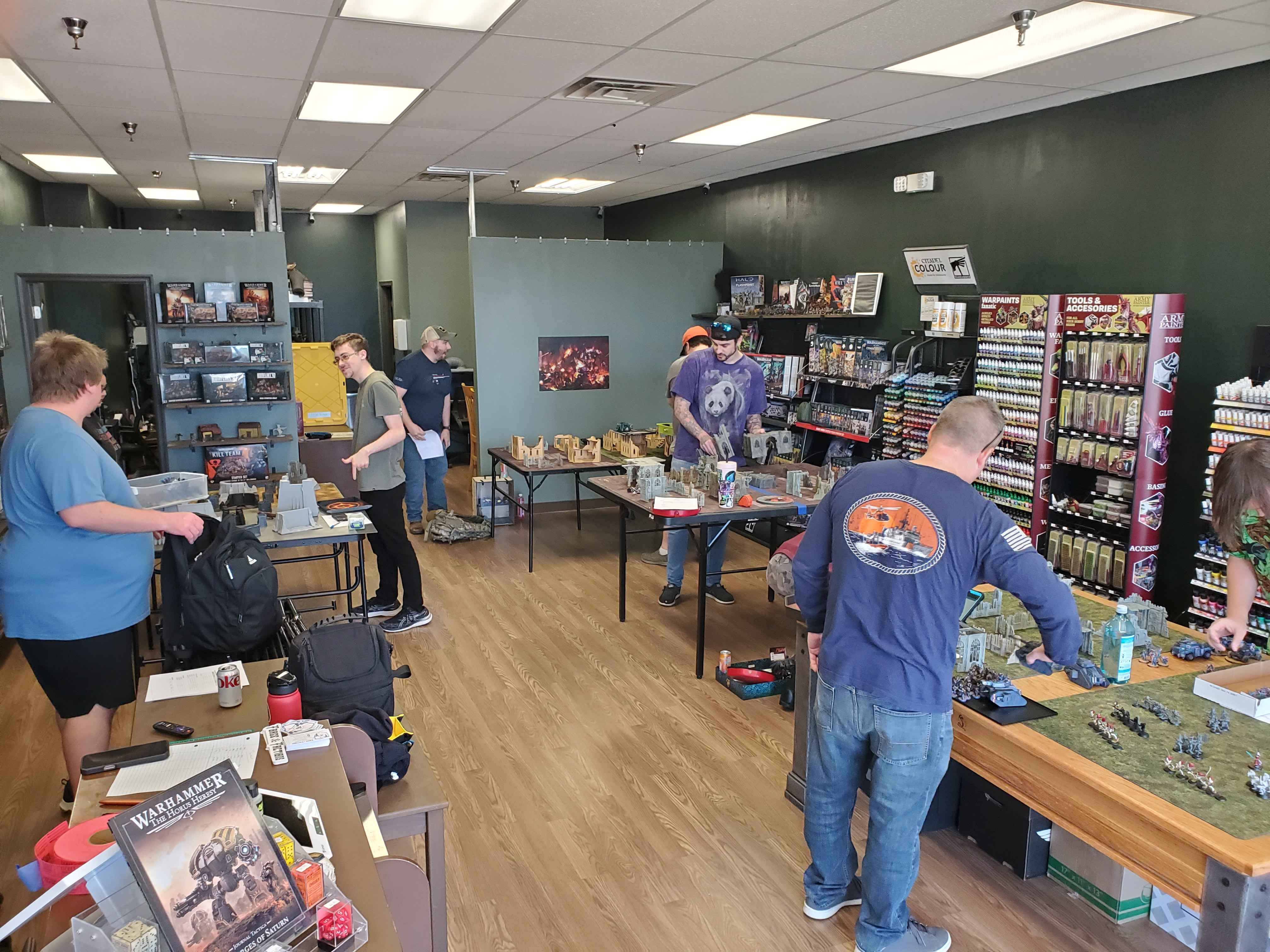 People shopping in a game store with shelves of products and a gaming table, some browsing and others working on miniatures.