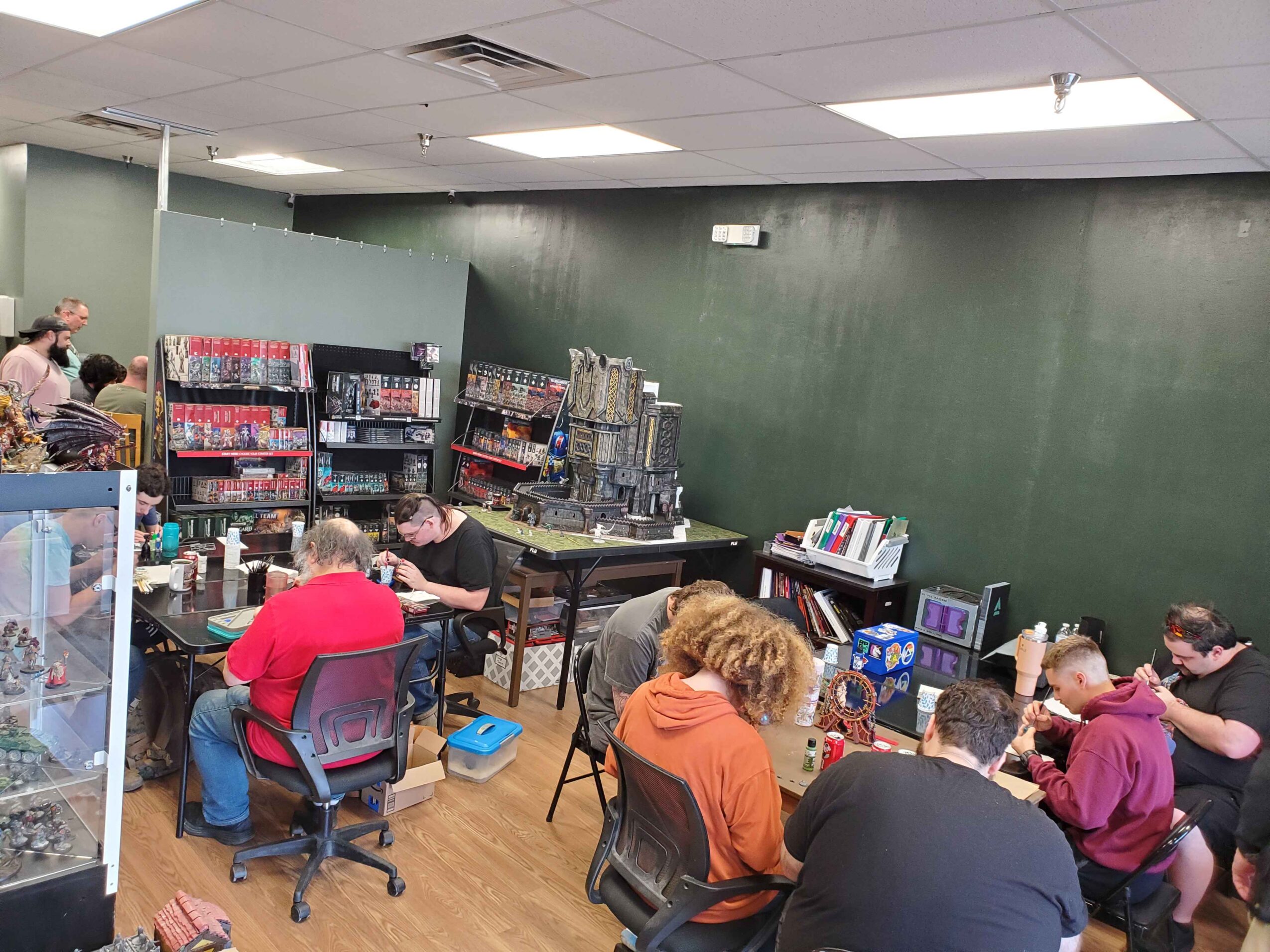 People sitting at tables in a store or classroom, with shelves of items and a green wall in the background.
