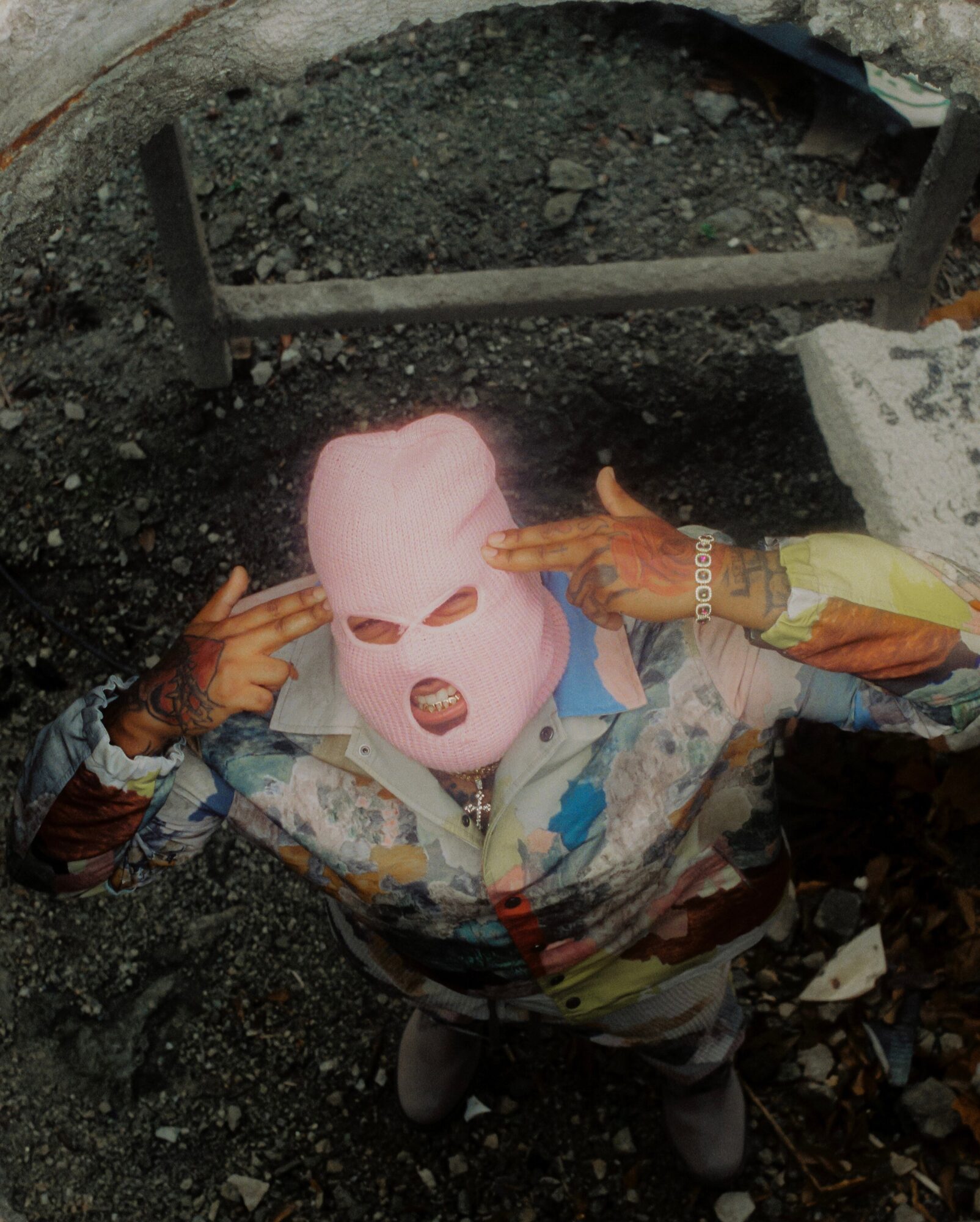 Person wearing a pink mask with eye and mouth holes, pointing fingers at their head, outdoors on gravel ground.