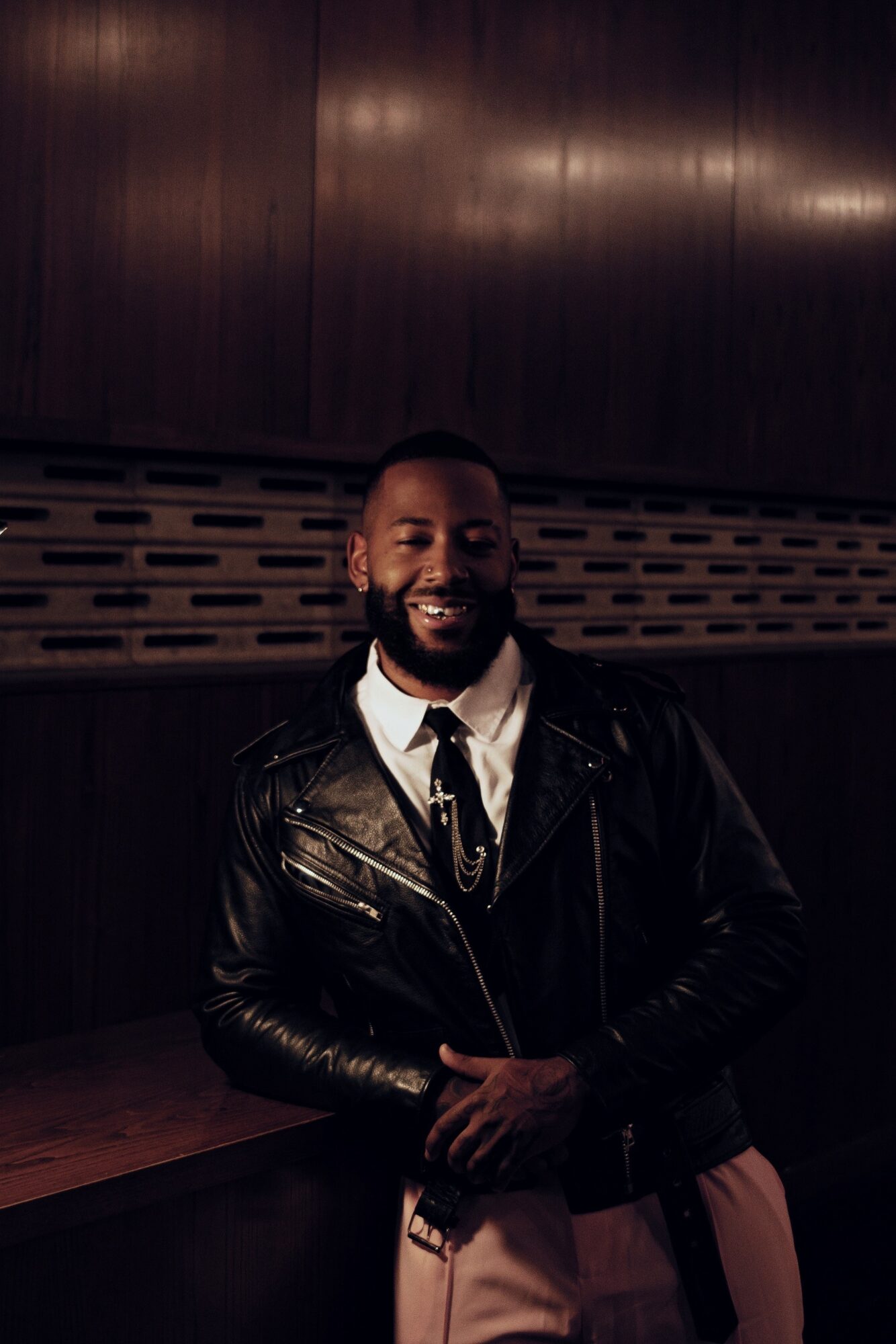 Man with a beard smiling, wearing a leather jacket and white shirt, standing indoors against a dark wooden wall.
