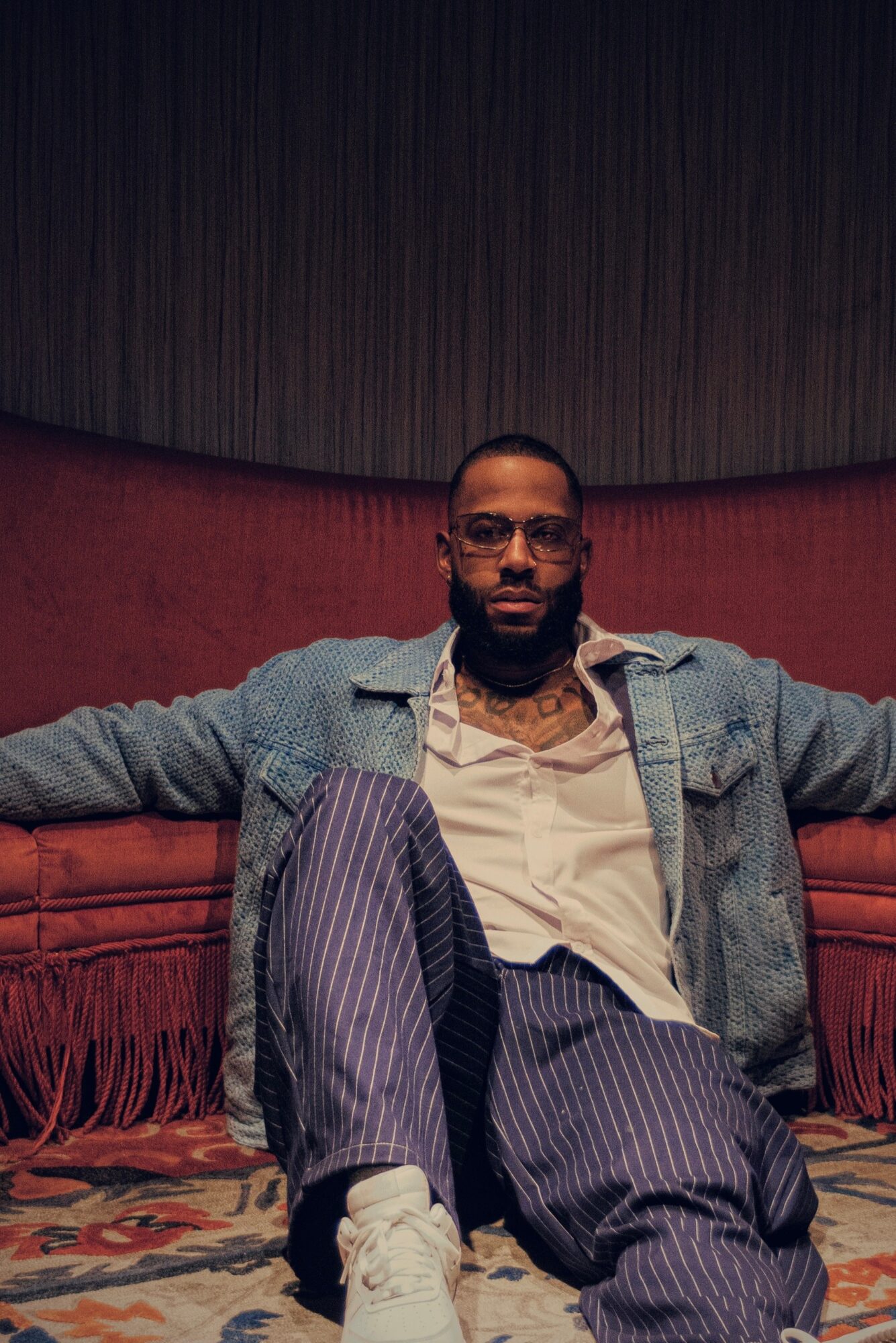 Man with glasses and beard sitting on a red cushioned bench with arms outstretched, wearing a gray jacket and striped pants.