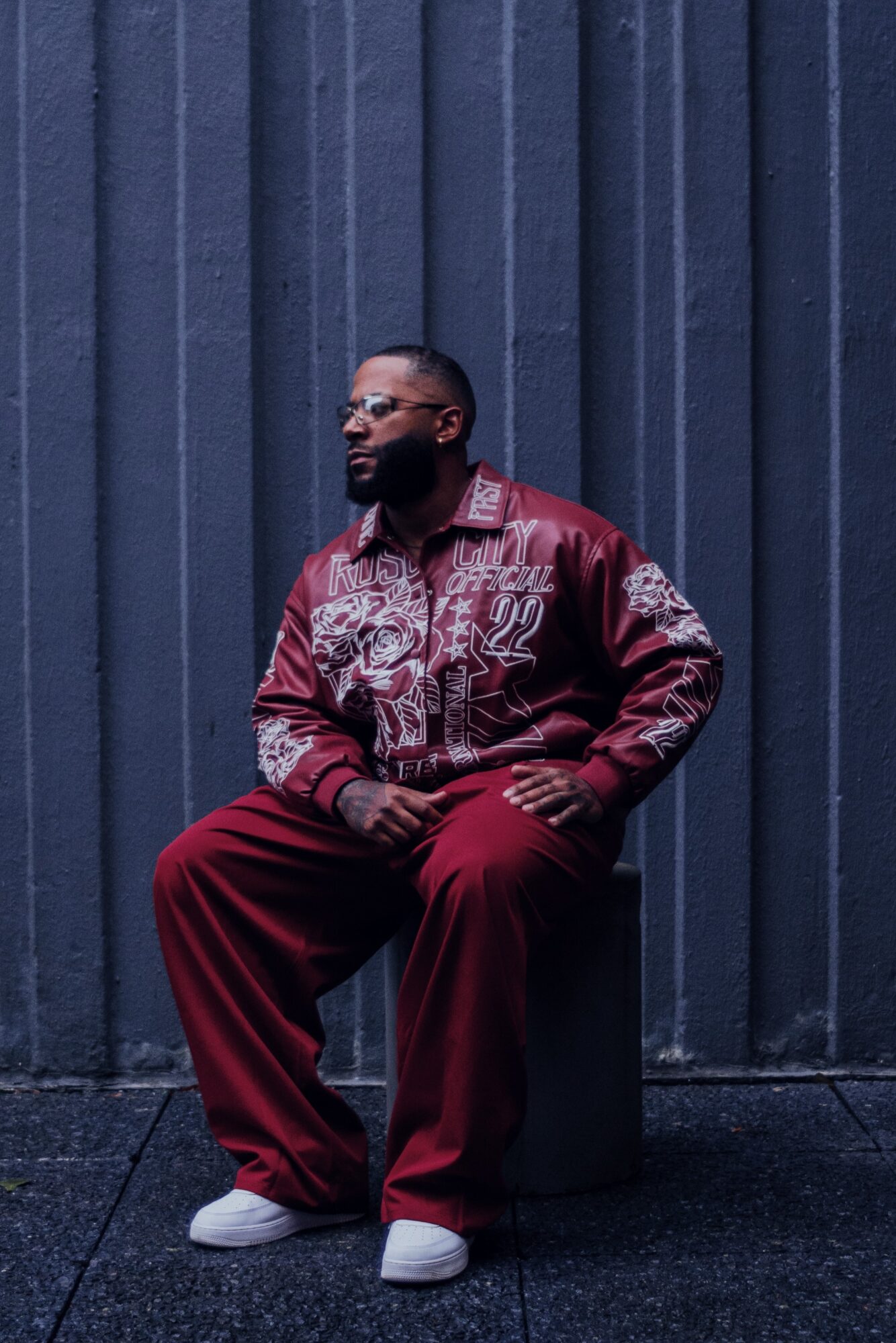 Man with glasses and beard sitting on a stool against a dark blue wall, wearing a patterned jacket and red pants.