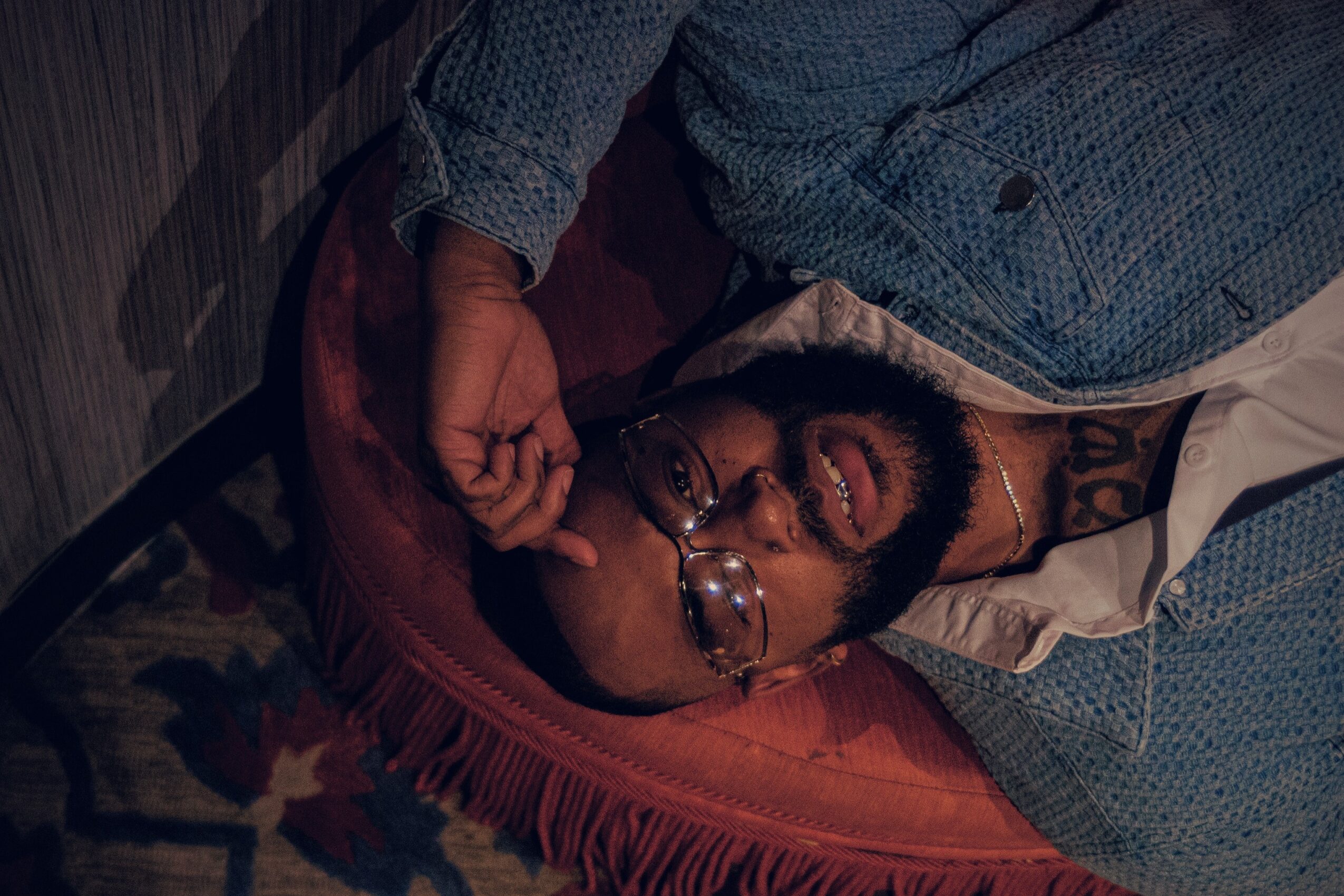 Person lying on a bed with glasses, beard, and open mouth, wearing a blue shirt, head on a pillow.