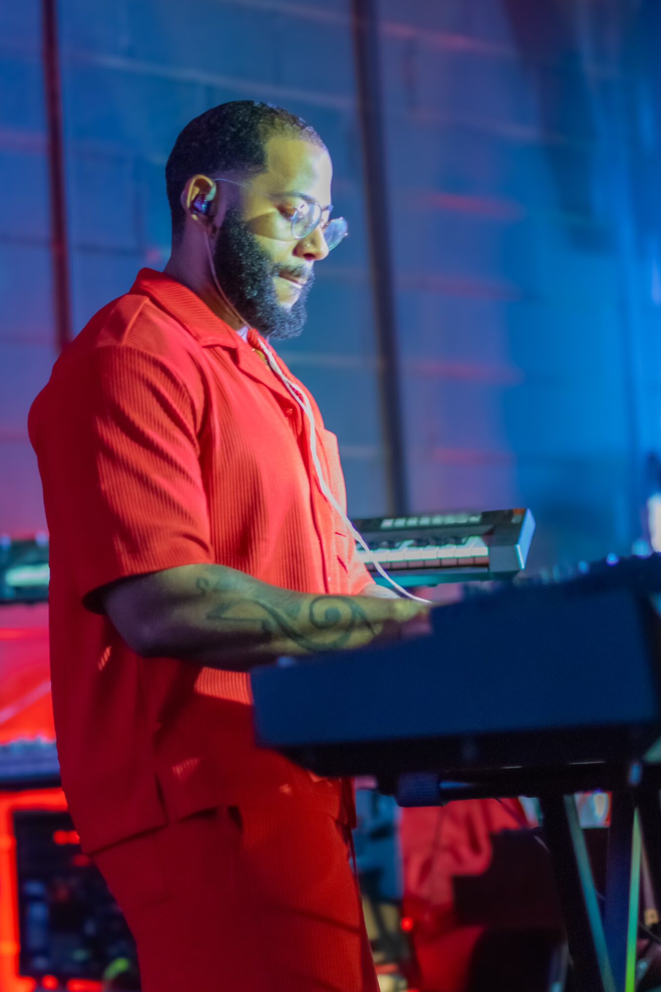 Person playing keyboard on stage, wearing red shirt, with colorful background lighting.