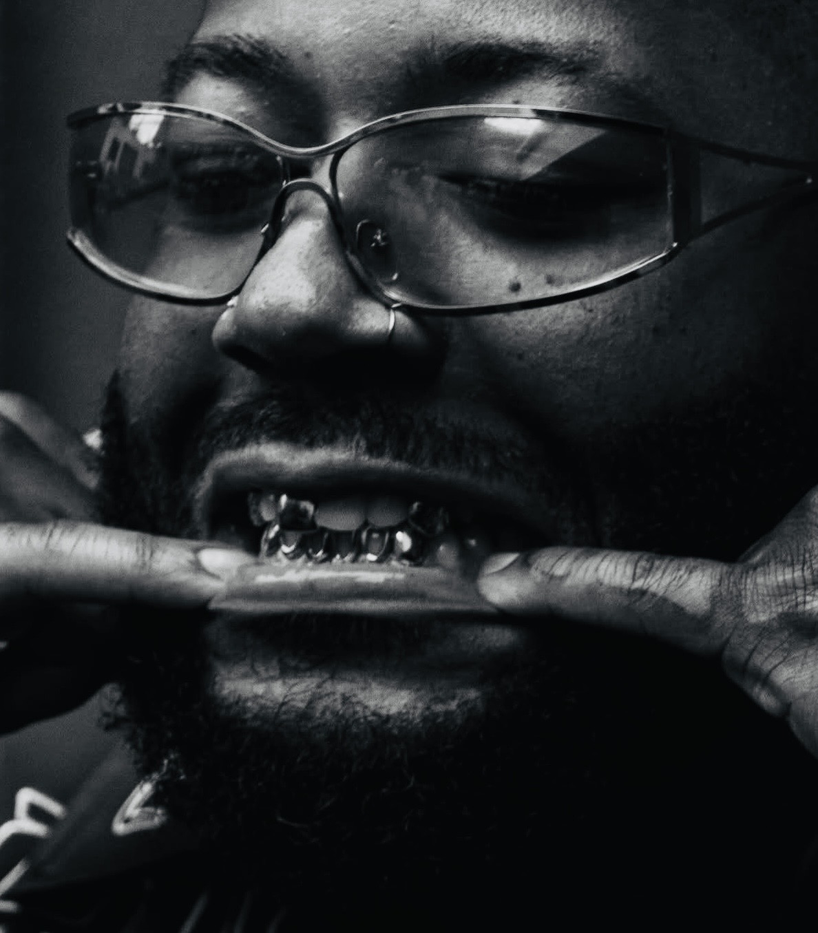 Close-up of a person with glasses showing their mouth with visible teeth, holding their fingers near lips, in black and white.