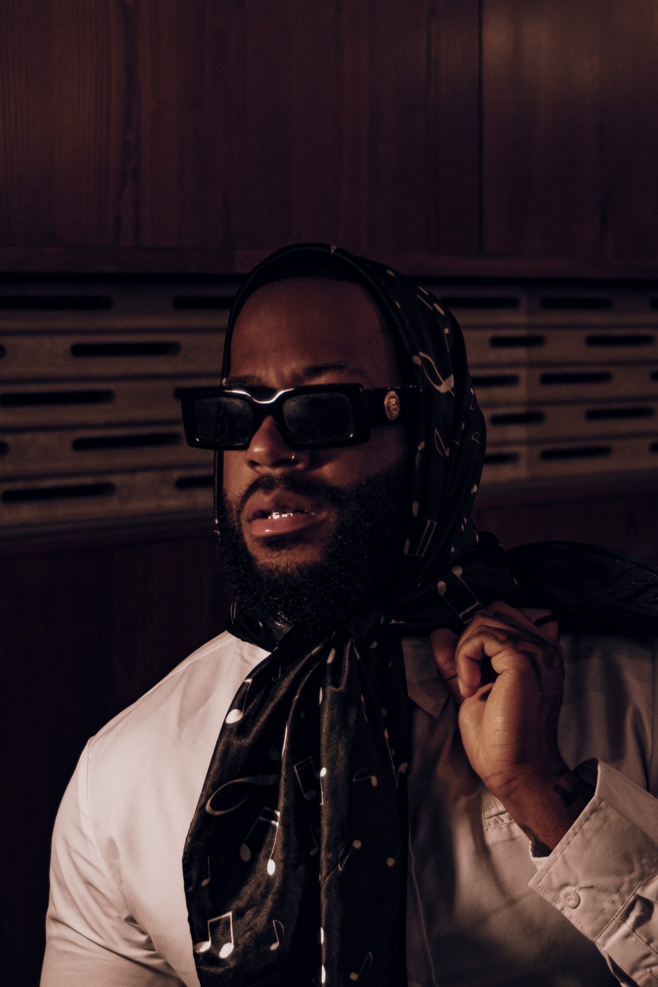Person wearing sunglasses and a headscarf, holding a strap, in a dimly lit room with wooden paneling.