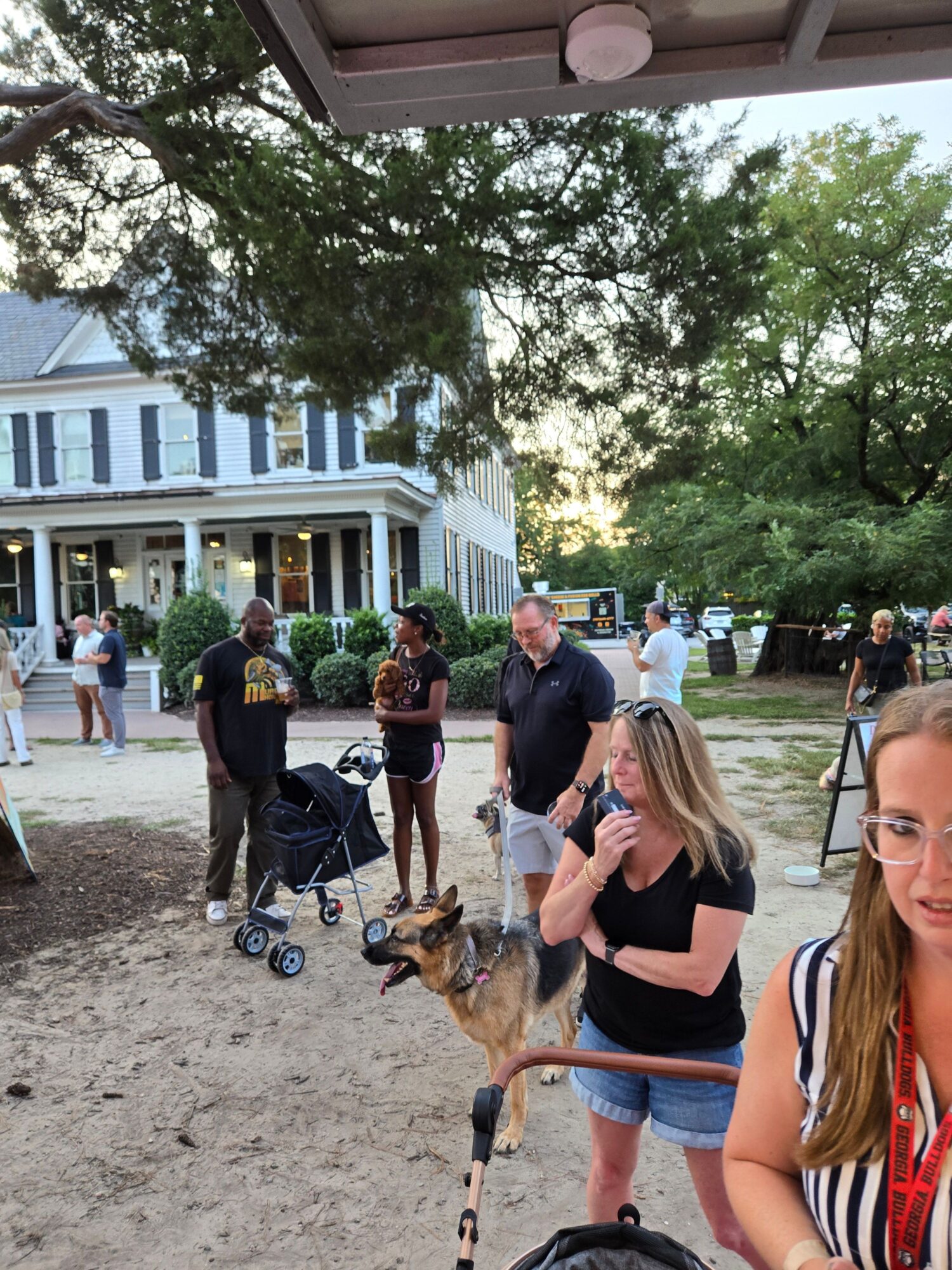 People walking and socializing outdoors near a building with trees and a dirt ground.