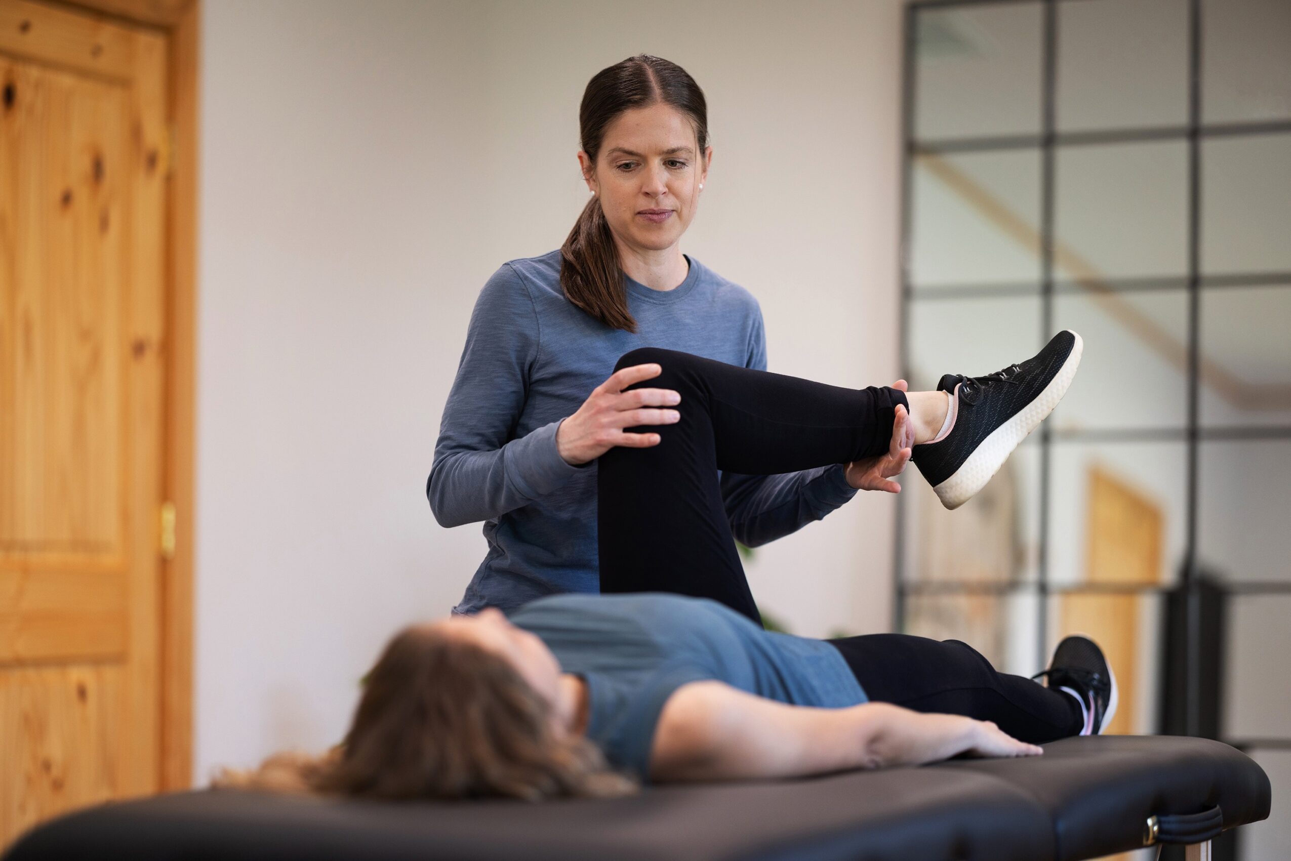 A woman in a blue sweater holds a person's leg while lying on a black massage table in a room with wooden and glass elements.